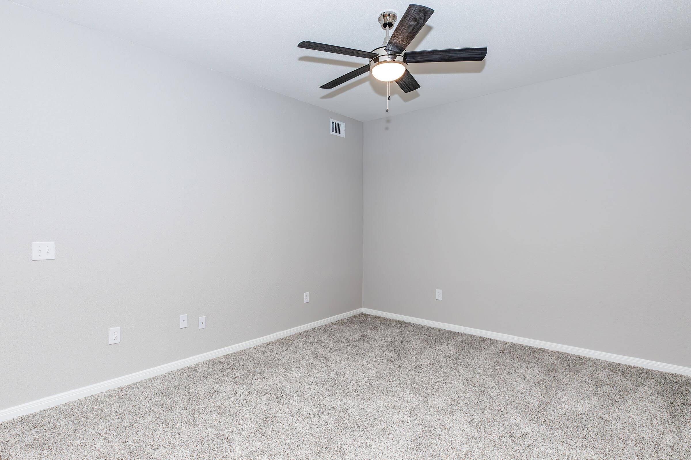 A spacious, empty room with light gray walls and a light-colored carpet. A ceiling fan with five blades hangs from the ceiling, providing a modern touch. There are multiple electrical outlets visible on the walls, and the overall ambiance is bright and clean, ready for decoration or furniture placement.