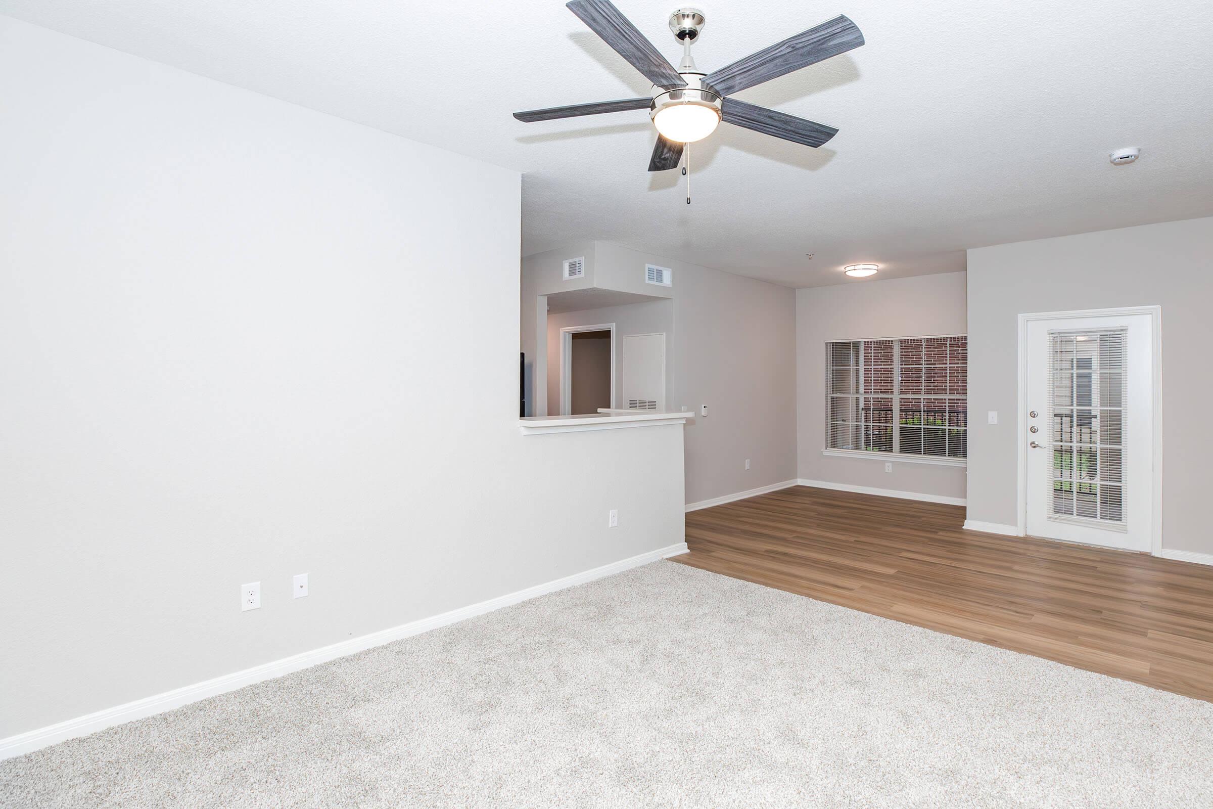 Spacious living room area with light-colored carpet and wood-style flooring. A ceiling fan is mounted on a smooth ceiling. There is a narrow ledge separating the room from an adjacent area. A large window with multiple panes and a glass door leads to a patio, allowing natural light to fill the space.