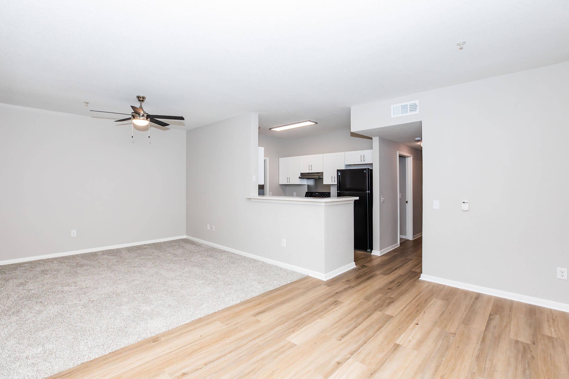 A spacious, modern living room featuring light-colored walls, a ceiling fan, and beige carpet. In the background, there is an open kitchen with white cabinetry and stainless steel appliances. The flooring transitions to light wood, creating a warm and inviting atmosphere.