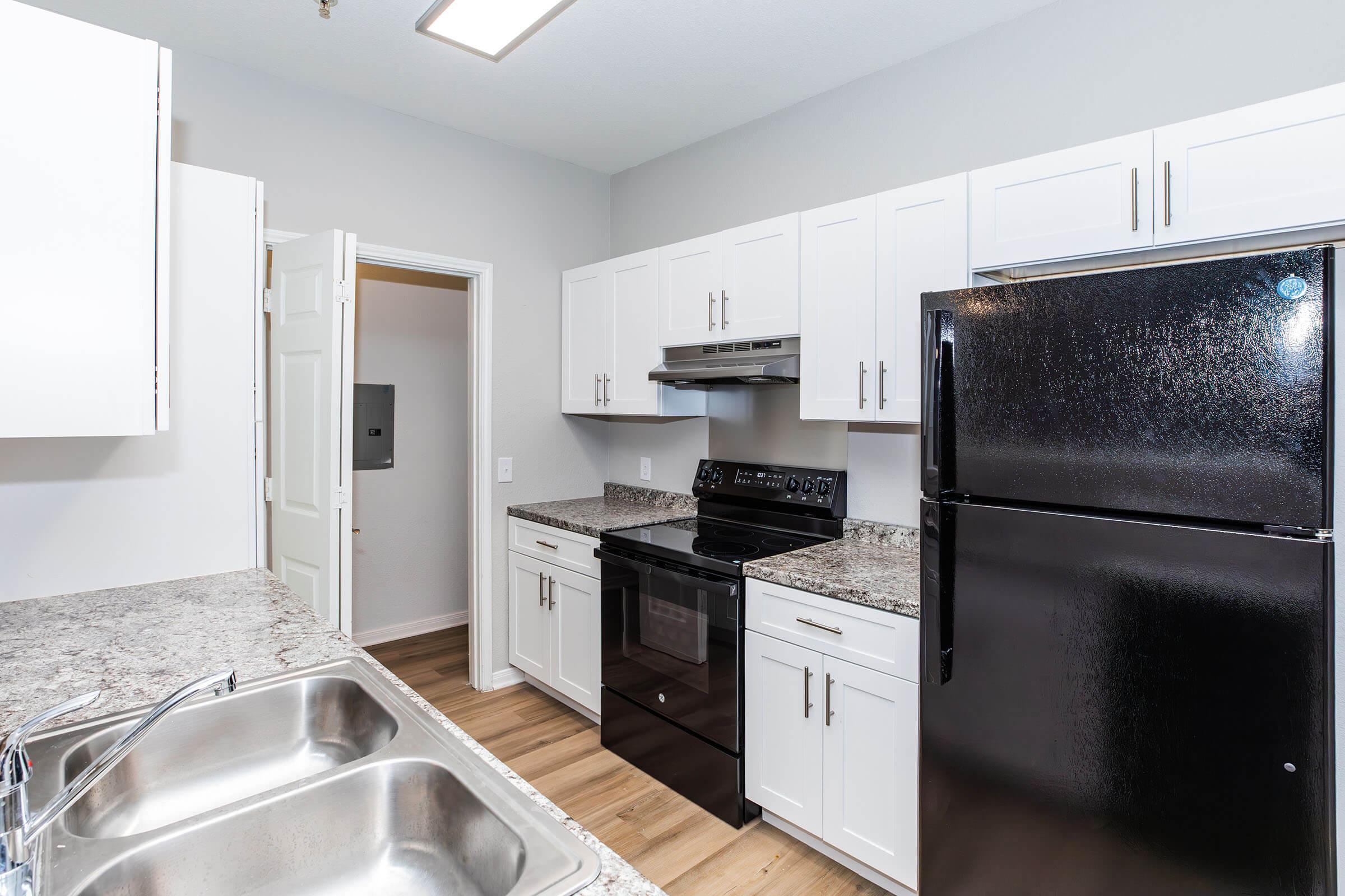 Modern kitchen featuring white cabinetry, granite countertops, and black appliances, including an oven and refrigerator. The space is well-lit with a ceiling light and has wood-like flooring. A doorway leads to another room, while a double sink is visible in the foreground.