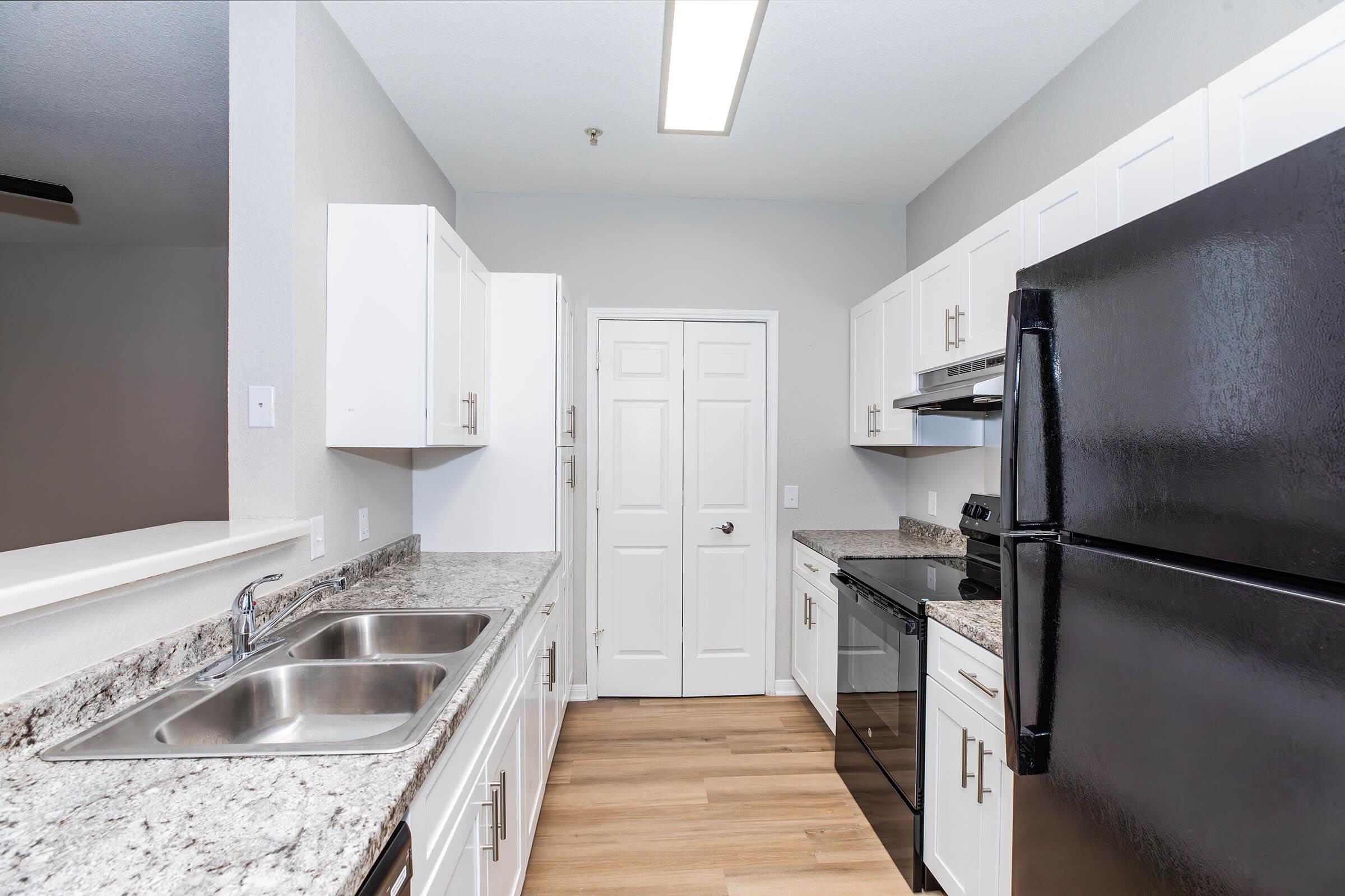 Modern kitchen featuring granite countertops, white cabinets, and stainless steel appliances. Includes a double sink, black refrigerator, and an oven with a sleek design. The walls are painted light gray, and there is a door leading to another area of the home. The flooring is wooden, adding warmth to the space.