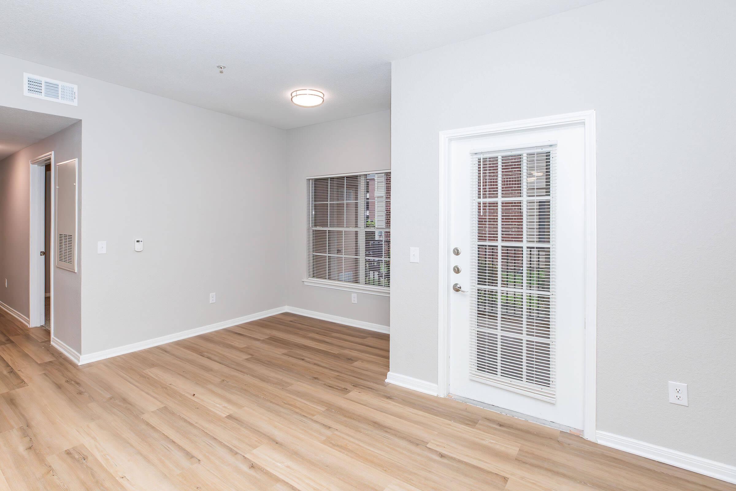 A bright, empty room with light gray walls and wooden flooring. There is a white door leading outside, a large window with blinds, and a small hallway visible in the background. The space appears modern and well-lit, ideal for furnishing and decoration.