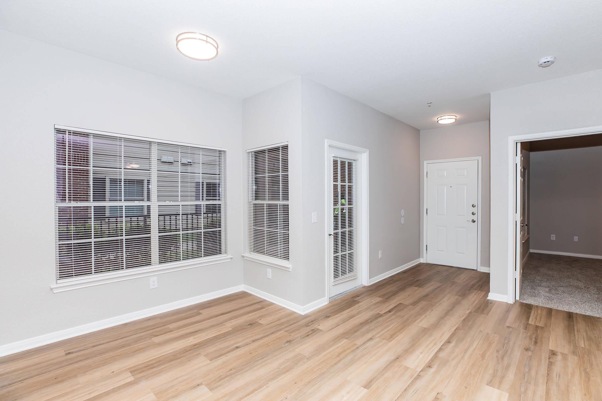 A bright, spacious room with light gray walls and wood-like laminate flooring. Large windows with blinds allow natural light, highlighting the open layout. There is a doorway leading to another room, and the entrance is visible on the right side. Modern ceiling lights provide additional illumination.