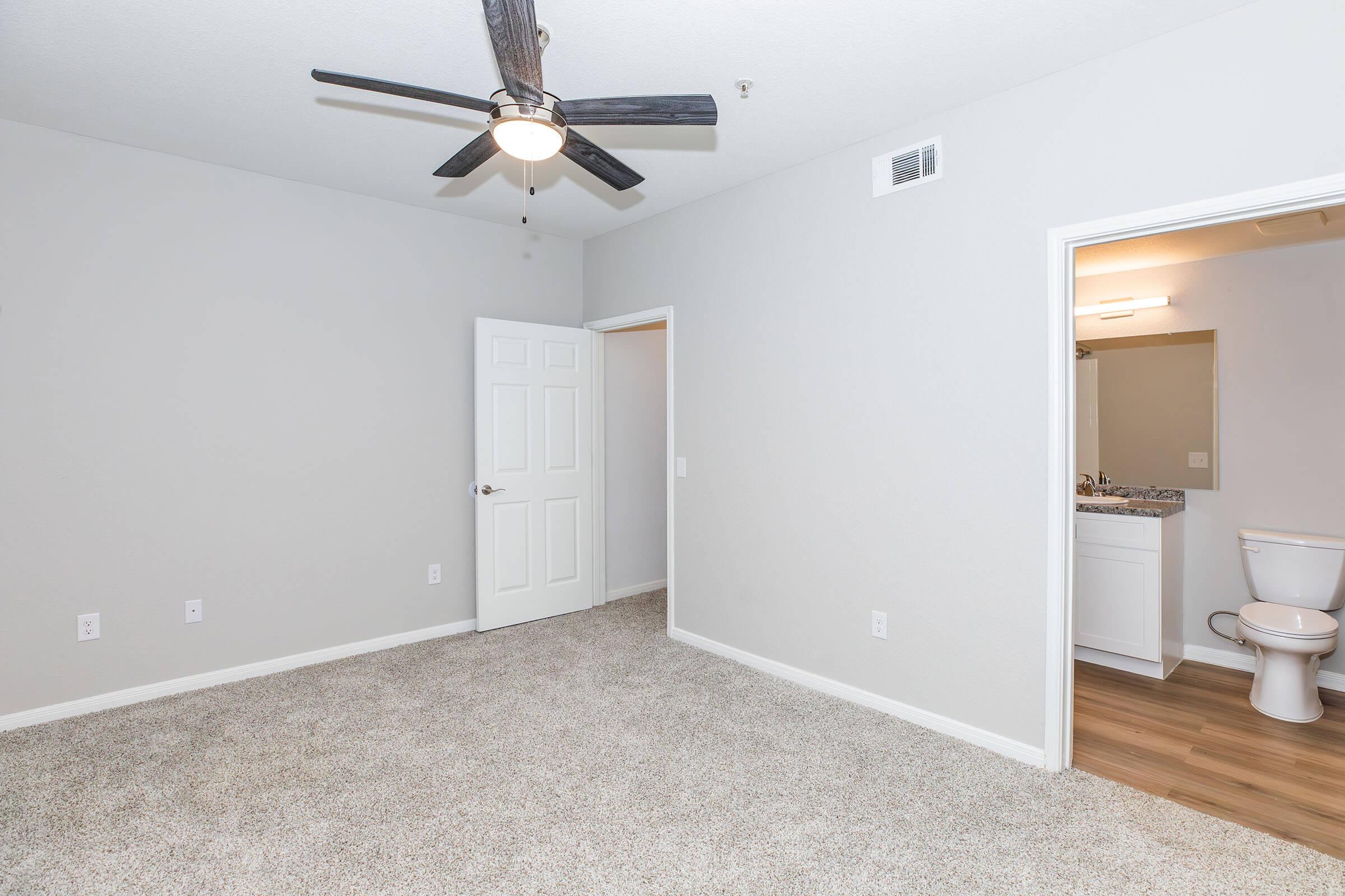 A spacious, light gray bedroom with a ceiling fan. There’s a doorway leading to a bathroom on the left and a closed door on the right. The floor is covered in soft, beige carpet, and the walls are plain without any decorations. The bathroom area includes a toilet visible through the open door.