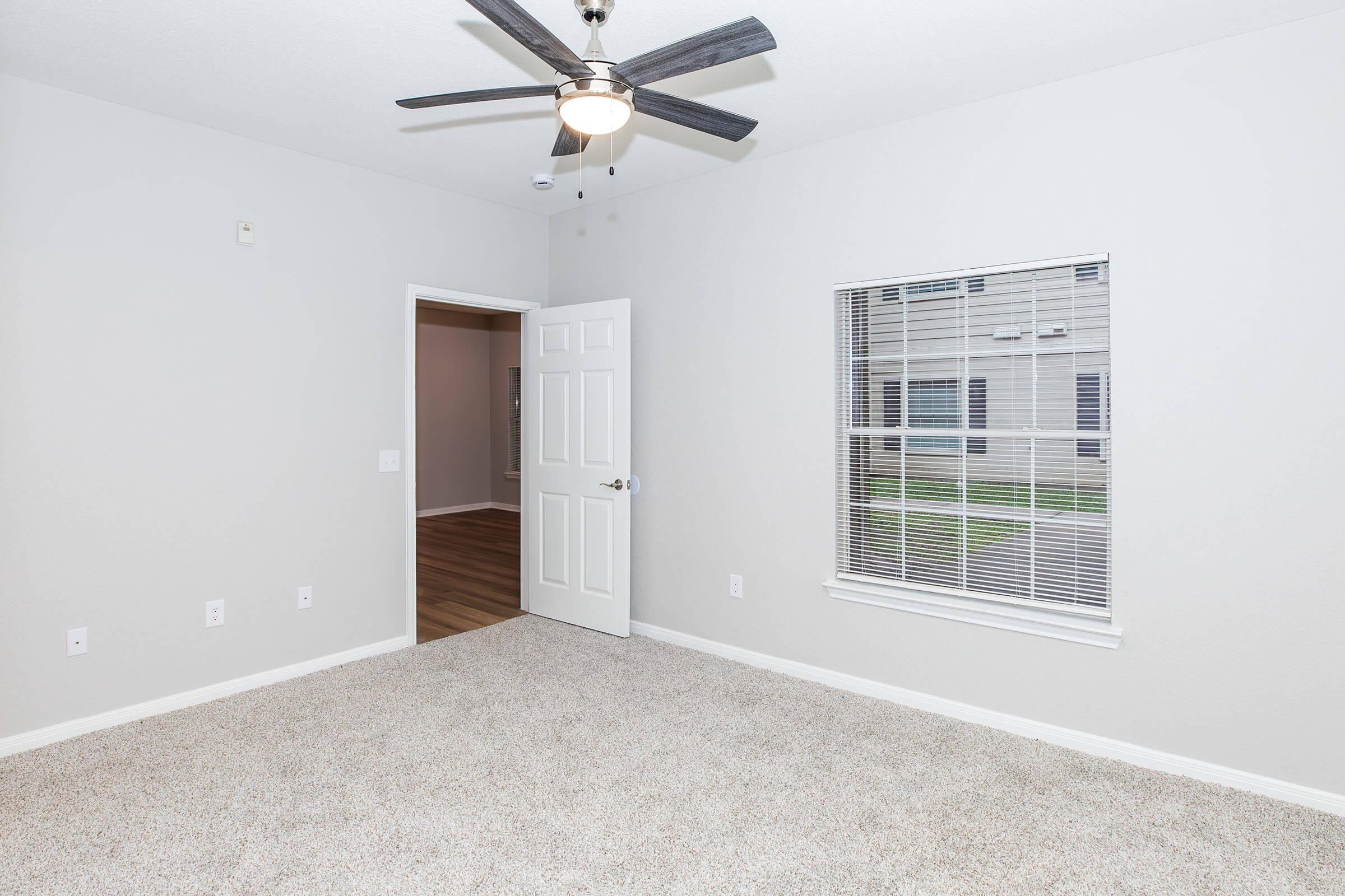 A light gray room with carpeted flooring, featuring a ceiling fan and a window with blinds. A door leads to another room, and the wall is free of decorations. Natural light streams in through the window, illuminating the space.