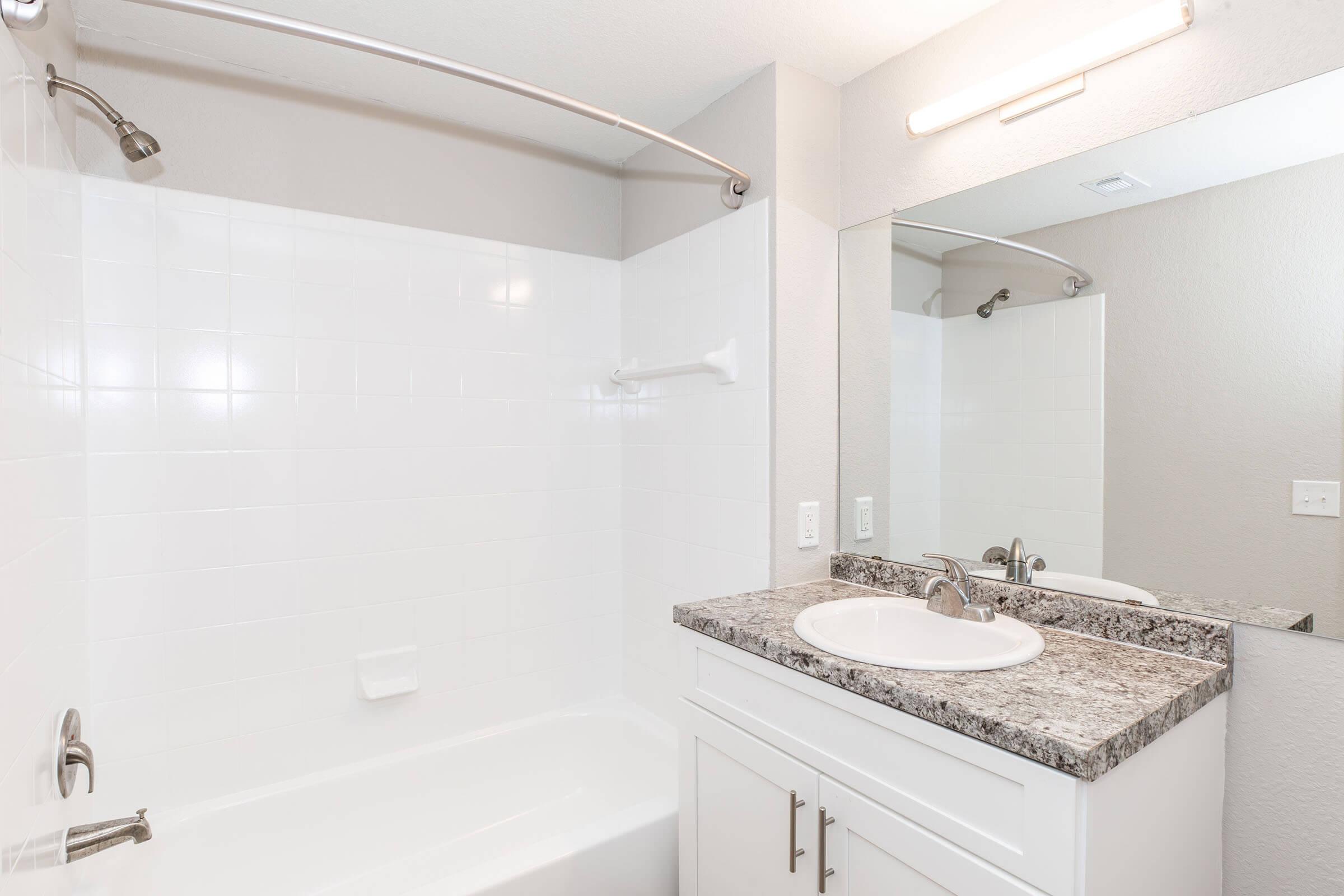 A clean and modern bathroom featuring a white tiled shower and bathtub, a granite countertop with a circular sink, and a large mirror above the sink. The walls are painted in a light color, and there is a single light fixture above the mirror, providing ample illumination. The space appears well-maintained and inviting.