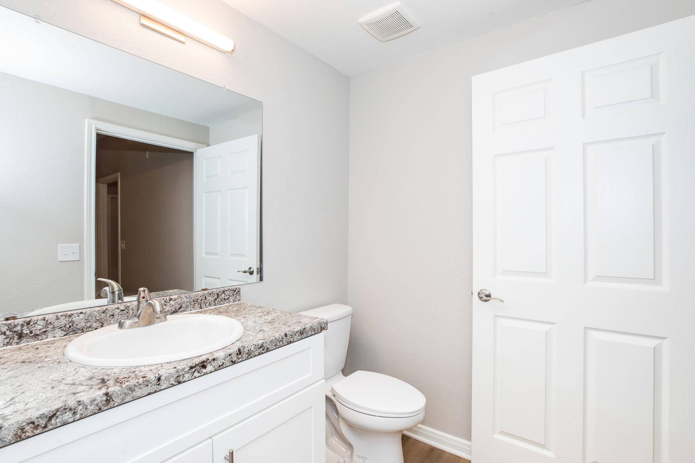 A clean and modern bathroom featuring a white sink with a granite countertop, a toilet, a large mirror, and light gray walls. A white door leads to another area, and there is a ceiling vent above the sink. The space is well-lit and organized, creating a fresh and inviting atmosphere.