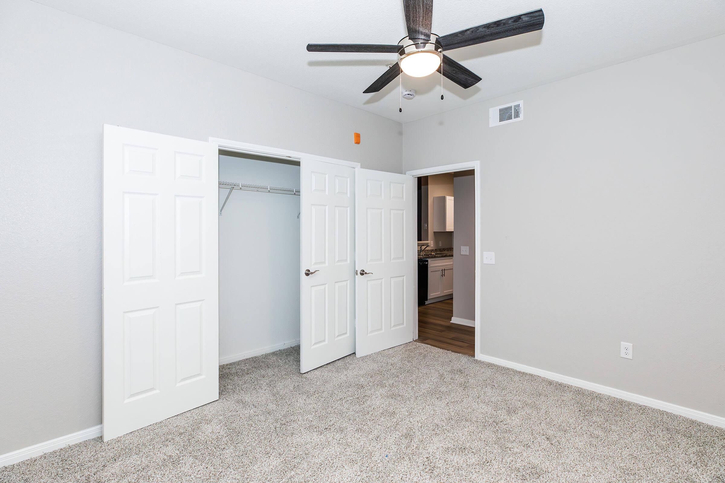 A well-lit bedroom with light gray walls and plush carpet. Two white double doors are open, revealing a closet space on the left. A door on the right leads to another room. The ceiling features a black ceiling fan with wooden blades, enhancing the modern and cozy atmosphere.