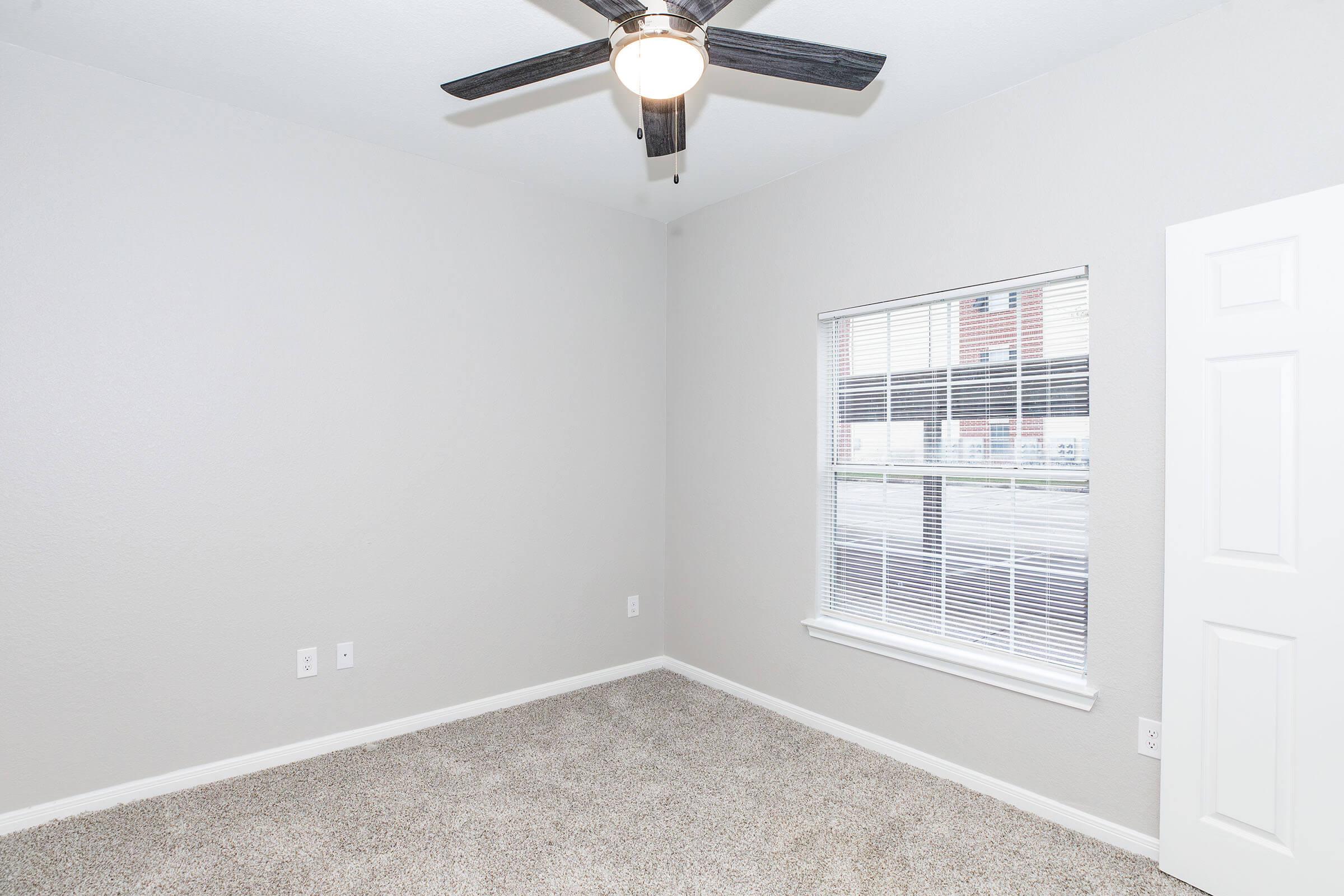 A clean, empty room featuring light gray walls and beige carpet. There is a ceiling fan with wooden blades, and a large window with white blinds, allowing natural light to enter. A white door is visible on the right side, leading to another space. The overall appearance is bright and airy.