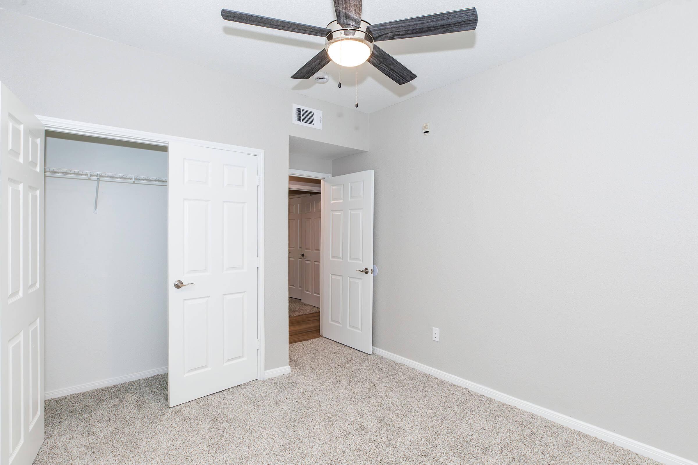 Empty room with light gray walls and beige carpet. A ceiling fan with black blades is mounted above. Two white doors are open, revealing a small closet and a doorway leading to another room. The space is well-lit and appears clean and fresh. Ideal for a living or bedroom area.