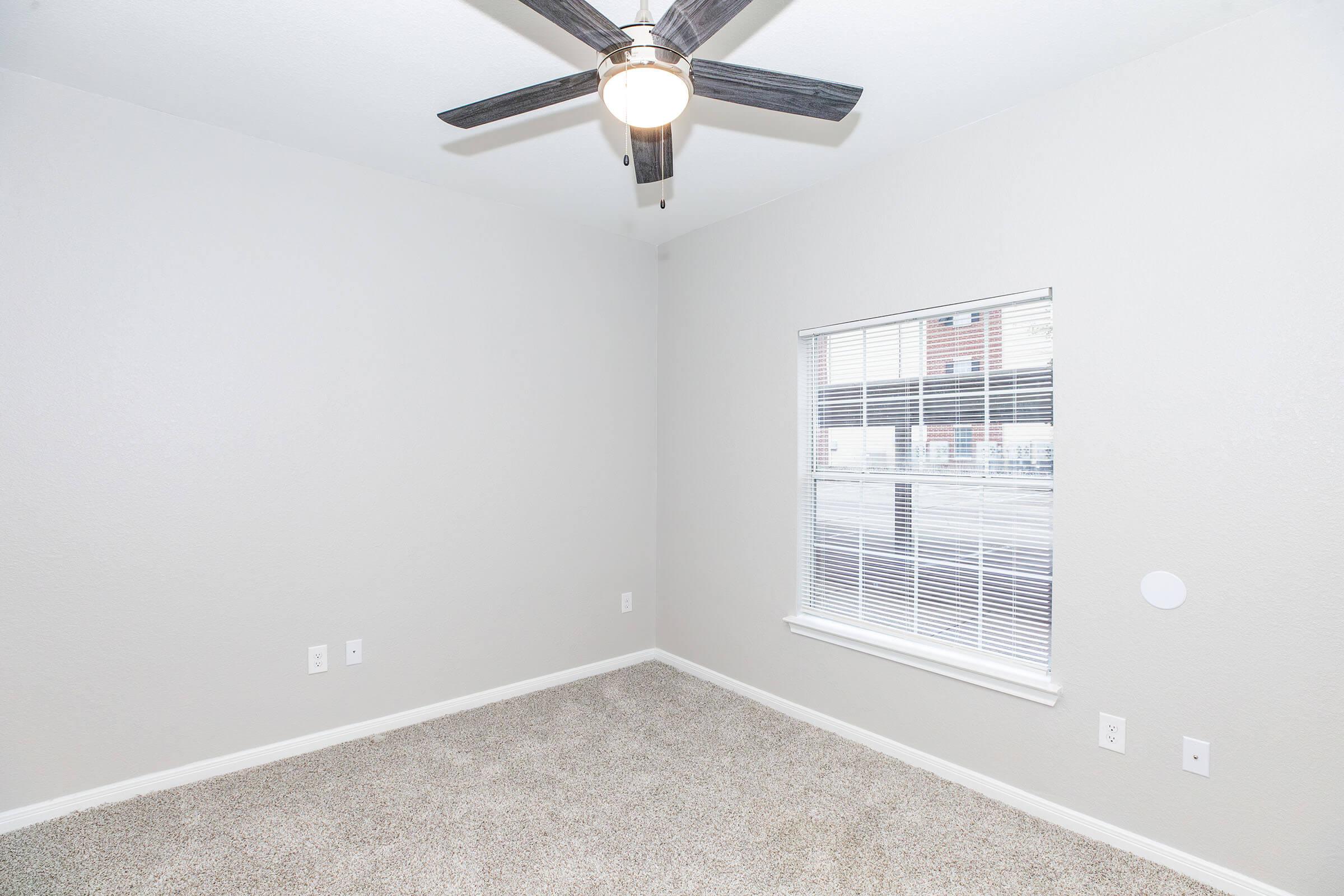 A sparsely furnished room featuring light gray walls and beige carpet. A ceiling fan with wooden blades hangs from the ceiling. There is a window with white blinds allowing natural light to enter the space. The room is empty and ready for furnishing.