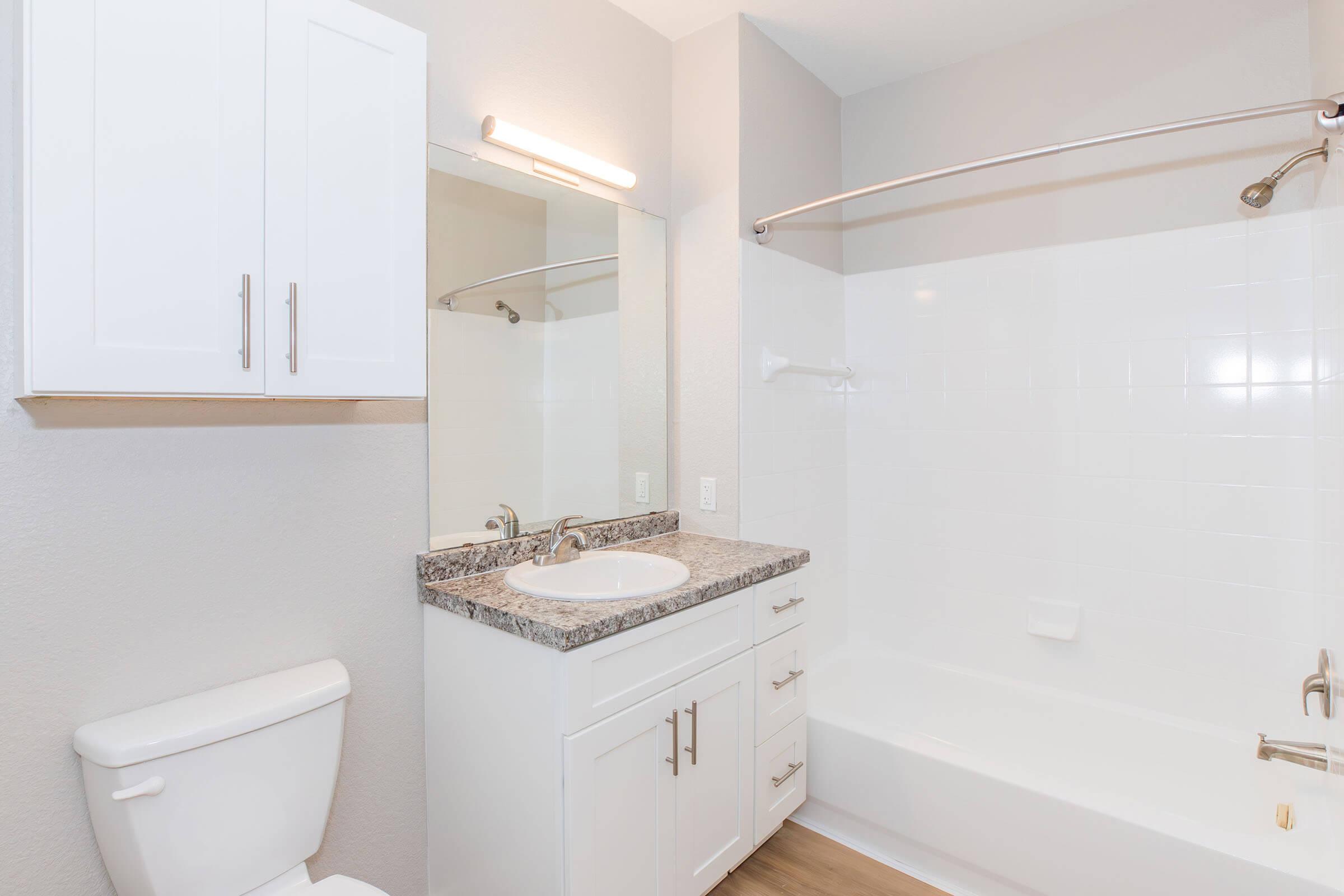 A modern bathroom featuring a white toilet, a vanity with a granite countertop and sink, and a bathtub/shower combo. There is a large mirror above the sink and neutral-colored walls. The flooring is a laminate wood finish, creating a clean and contemporary look.