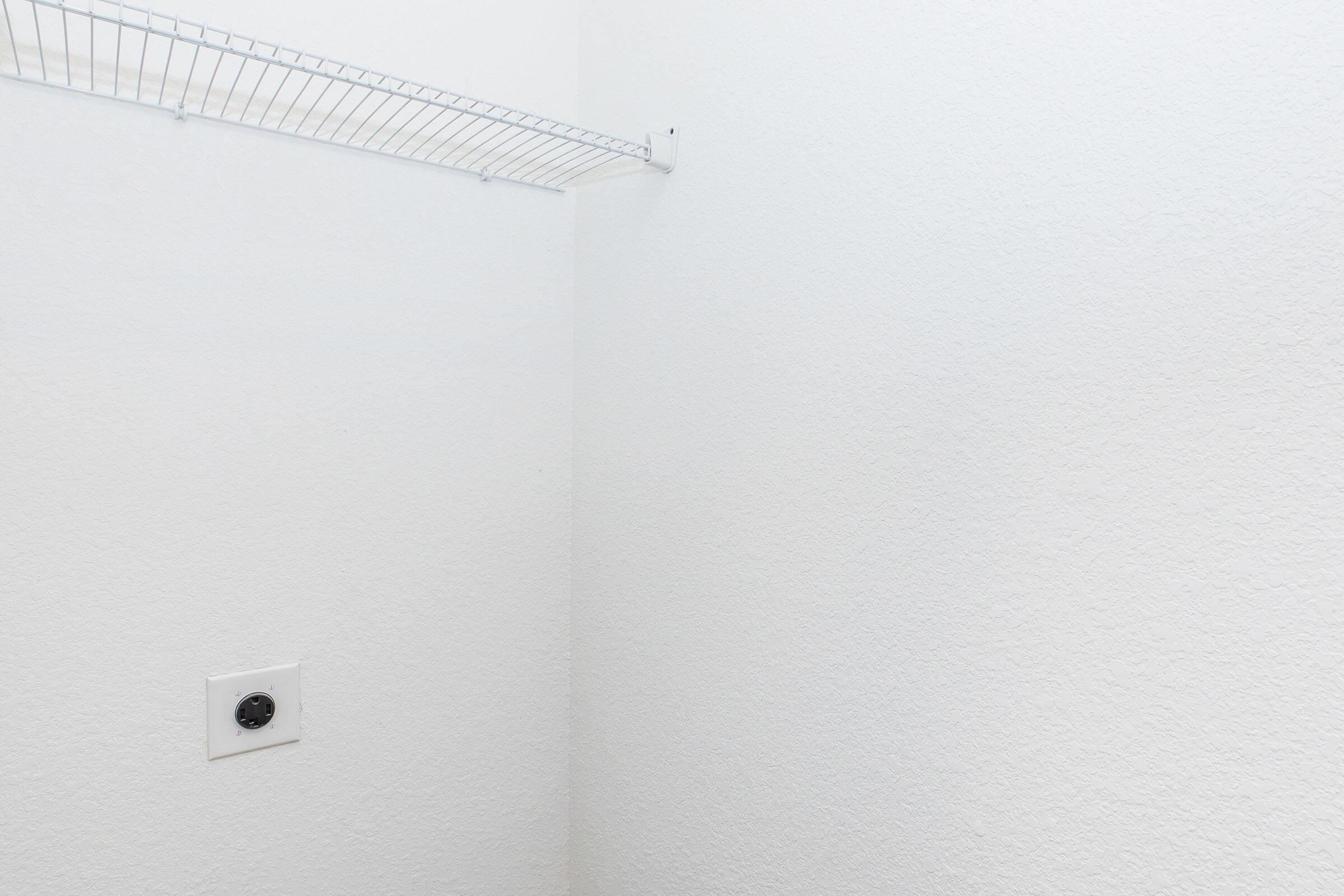 A small, empty closet with white walls. There is a wire shelf installed along the top, and a power outlet is visible on the wall. The overall look is clean and minimalistic, with no additional items or furniture present.