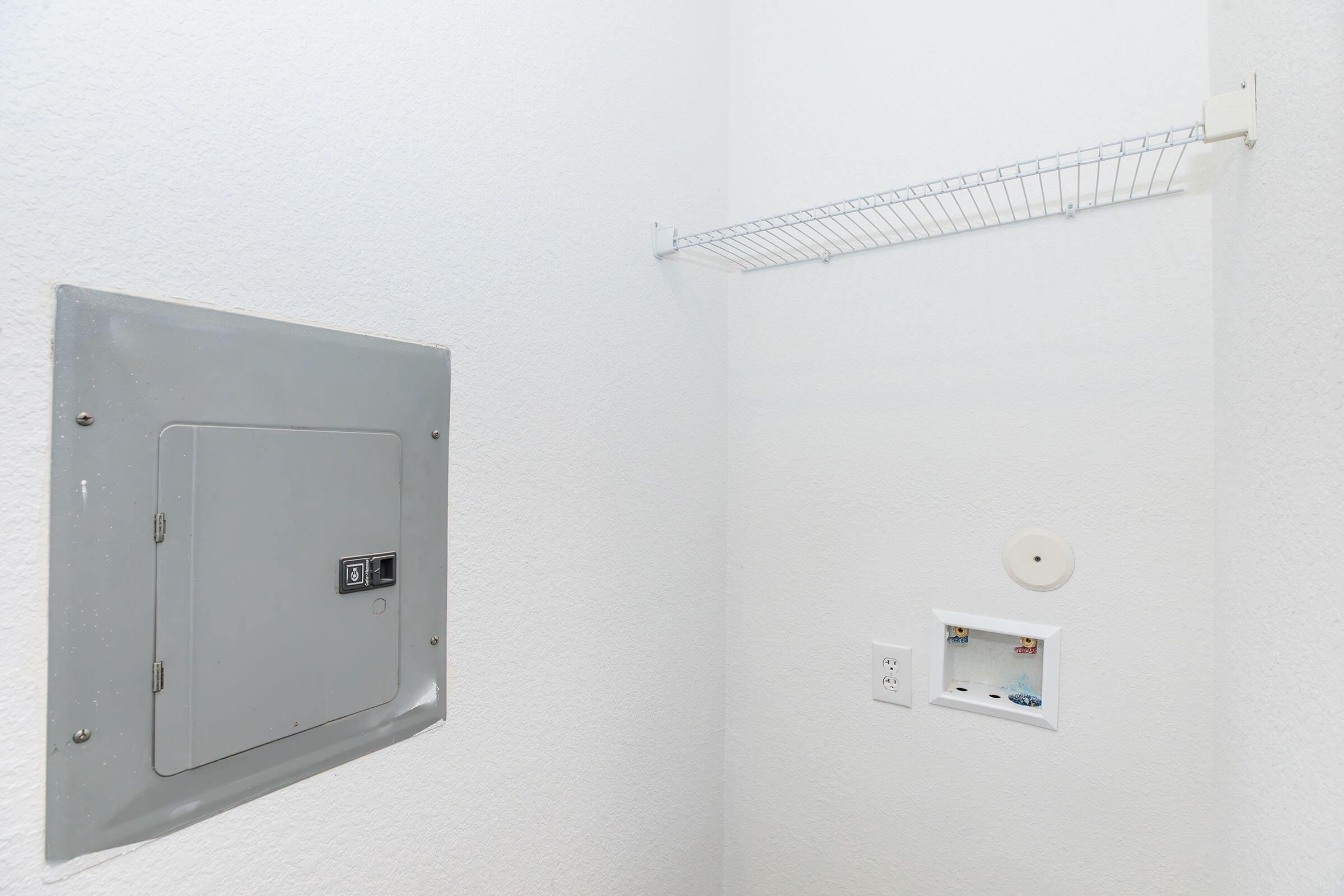 A small, empty laundry room featuring a gray electrical panel mounted on the wall, a white wire shelf above, and an electrical outlet with a circular cover. The walls are painted white, creating a clean and simple appearance.