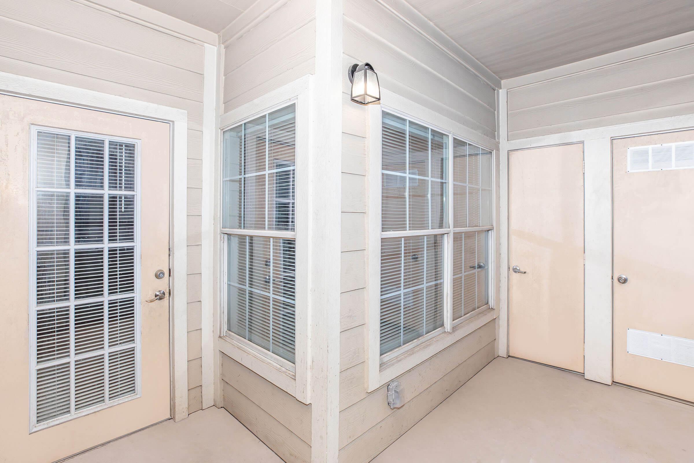 A corner view of a light-colored hallway featuring a door with a grid-patterned window and two adjacent doors. The walls are painted in a neutral tone, and there are windows with blinds allowing natural light to enter. A small wall-mounted light fixture is visible near the ceiling.