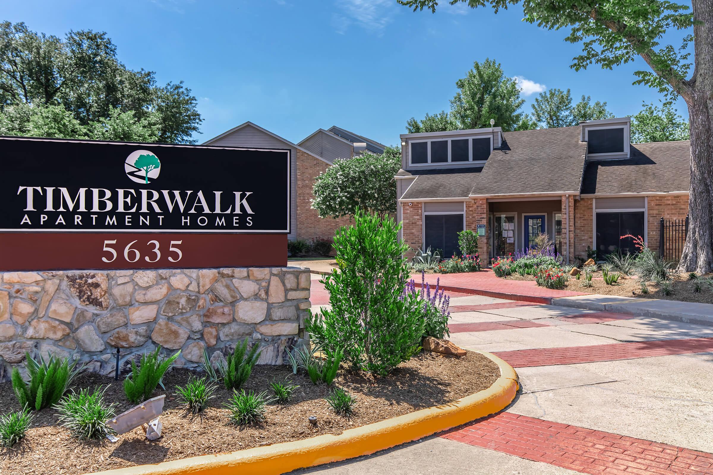 Timberwalk Apartment Homes sign in front of a landscaped building with stone accents. The pathway is lined with greenery and flowers, creating a welcoming atmosphere. Clear blue sky and trees visible in the background.