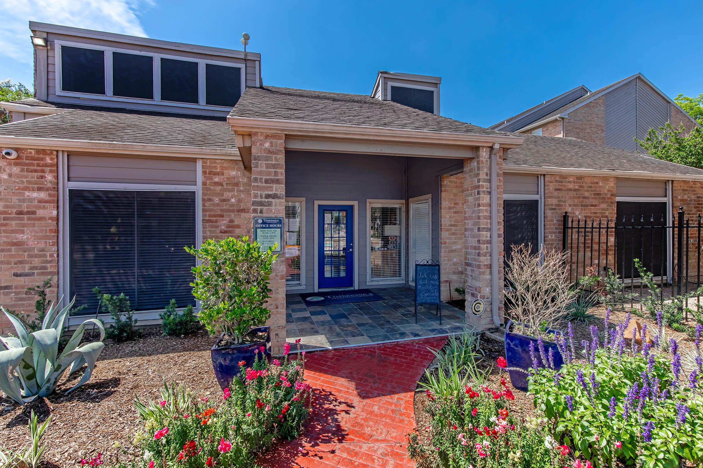 A welcoming building entrance with a blue door framed by brick walls. The pathway is lined with colorful flowers and plants, alongside decorative pots. Bright blue skies overhead and a well-maintained garden enhance the inviting atmosphere.