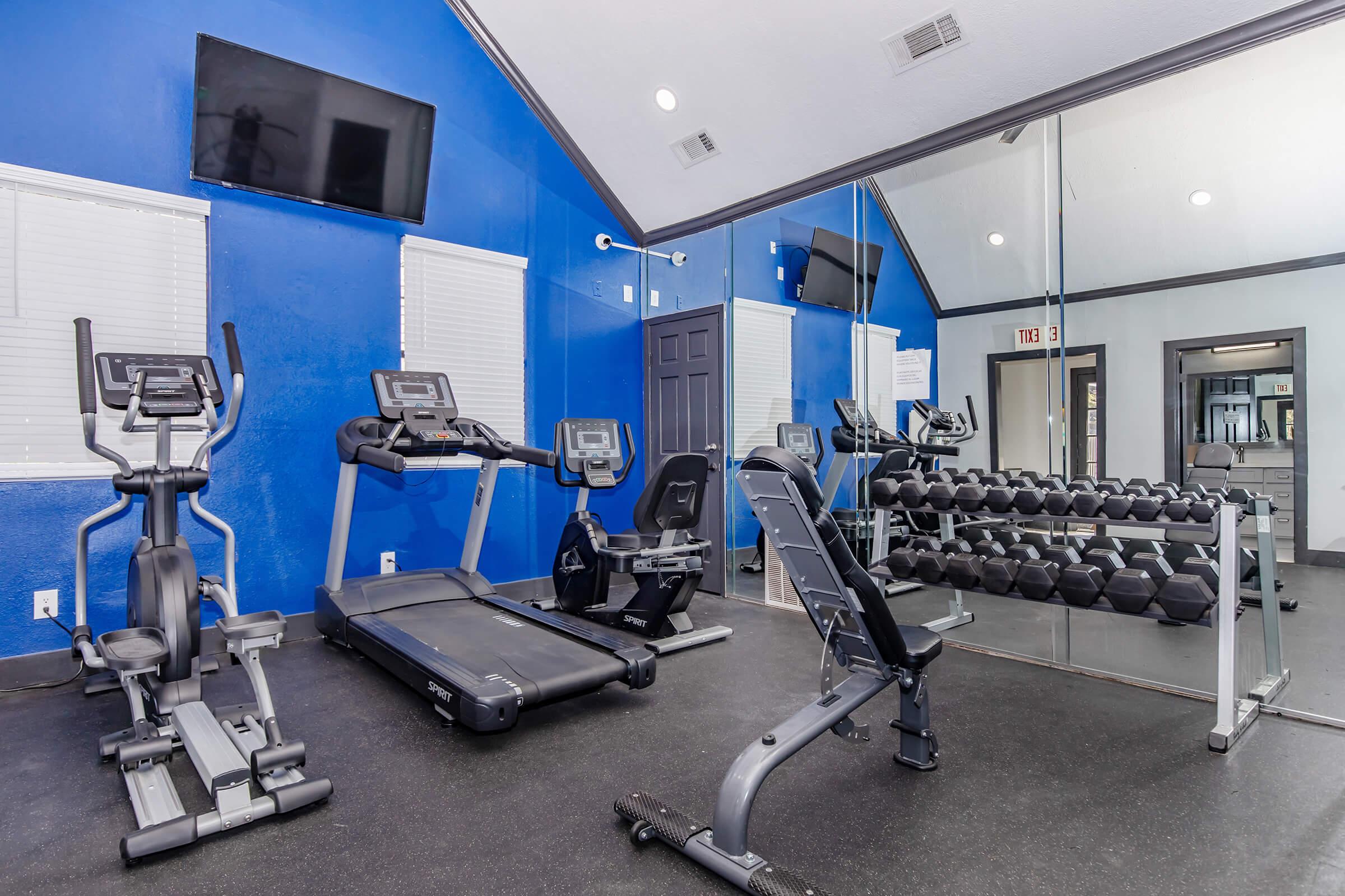 A well-equipped gym featuring elliptical machines, a treadmill, and a weight bench with dumbbells. The walls are painted blue, and there are mirrors and flat-screen TVs mounted on the walls, creating a modern workout environment. Bright lighting enhances the space, making it inviting for fitness activities.