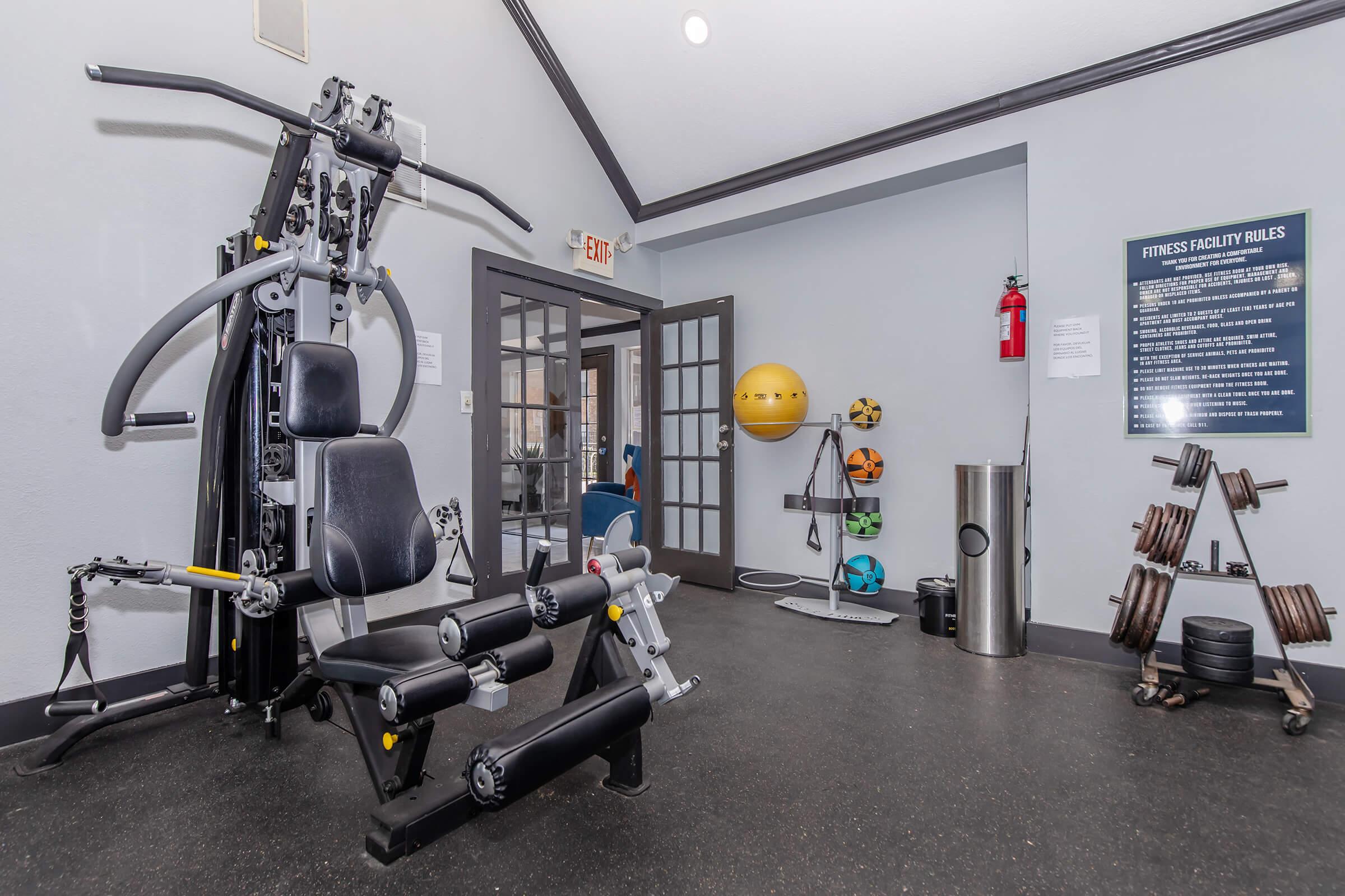 A spacious fitness room featuring a multi-gym machine, a set of free weights, a large stability ball, and various exercise equipment. The walls are painted in light colors, and fitness facility rules are displayed on a board. The area is well-lit with natural light coming through the door.