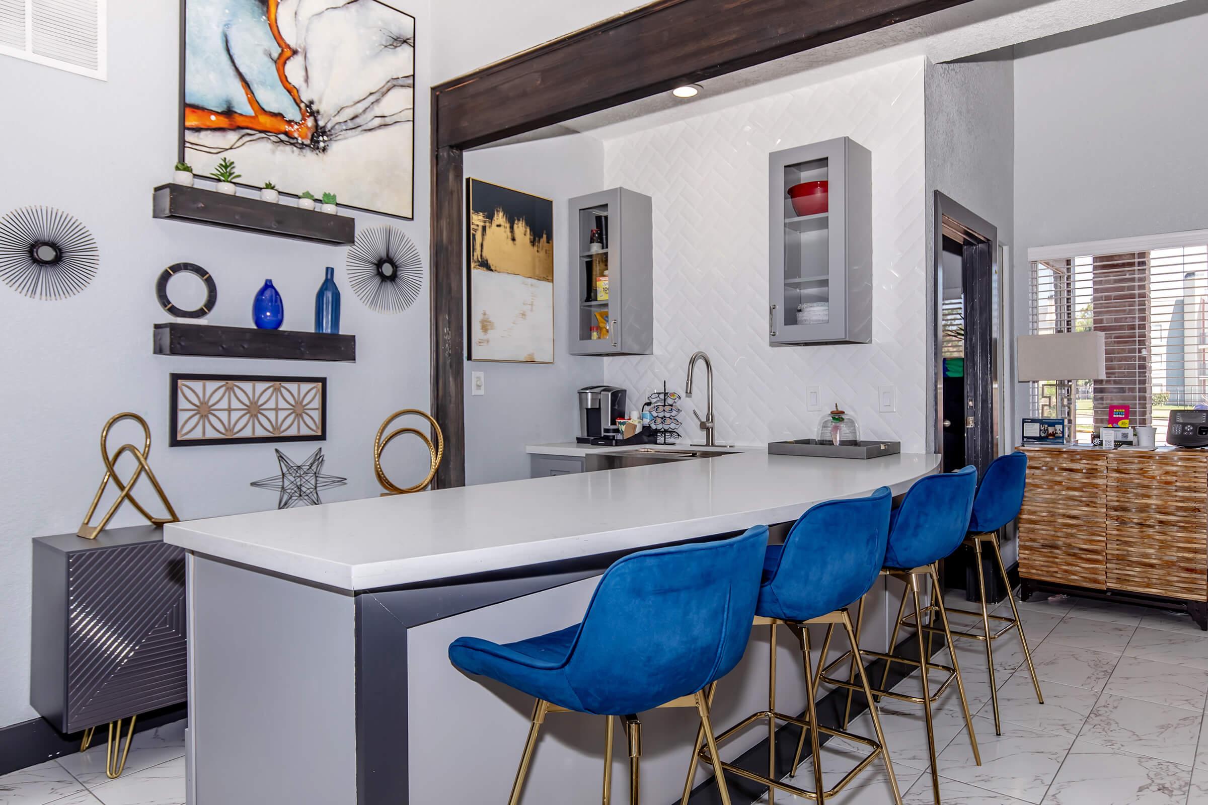 Modern kitchen featuring a sleek countertop with blue barstools, decorative shelves with various items, and contemporary wall art. The background includes a window with natural light and additional furniture, creating a stylish and inviting ambiance.