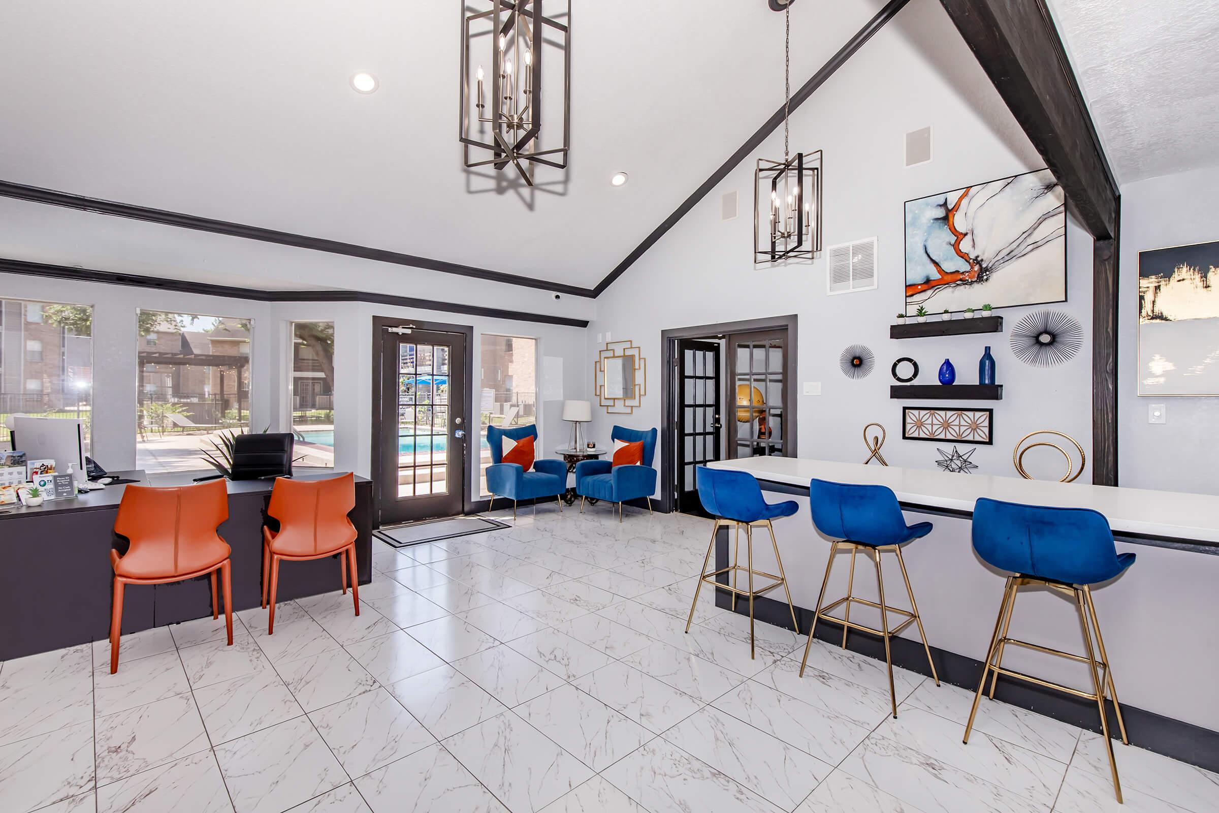 A modern lobby featuring a bar with high stools in blue and gold, orange and blue accent chairs, and large windows overlooking a pool area. The room has stylish light fixtures and contemporary decor, including artwork on the walls and a reception desk with a computer setup. Bright and welcoming atmosphere.