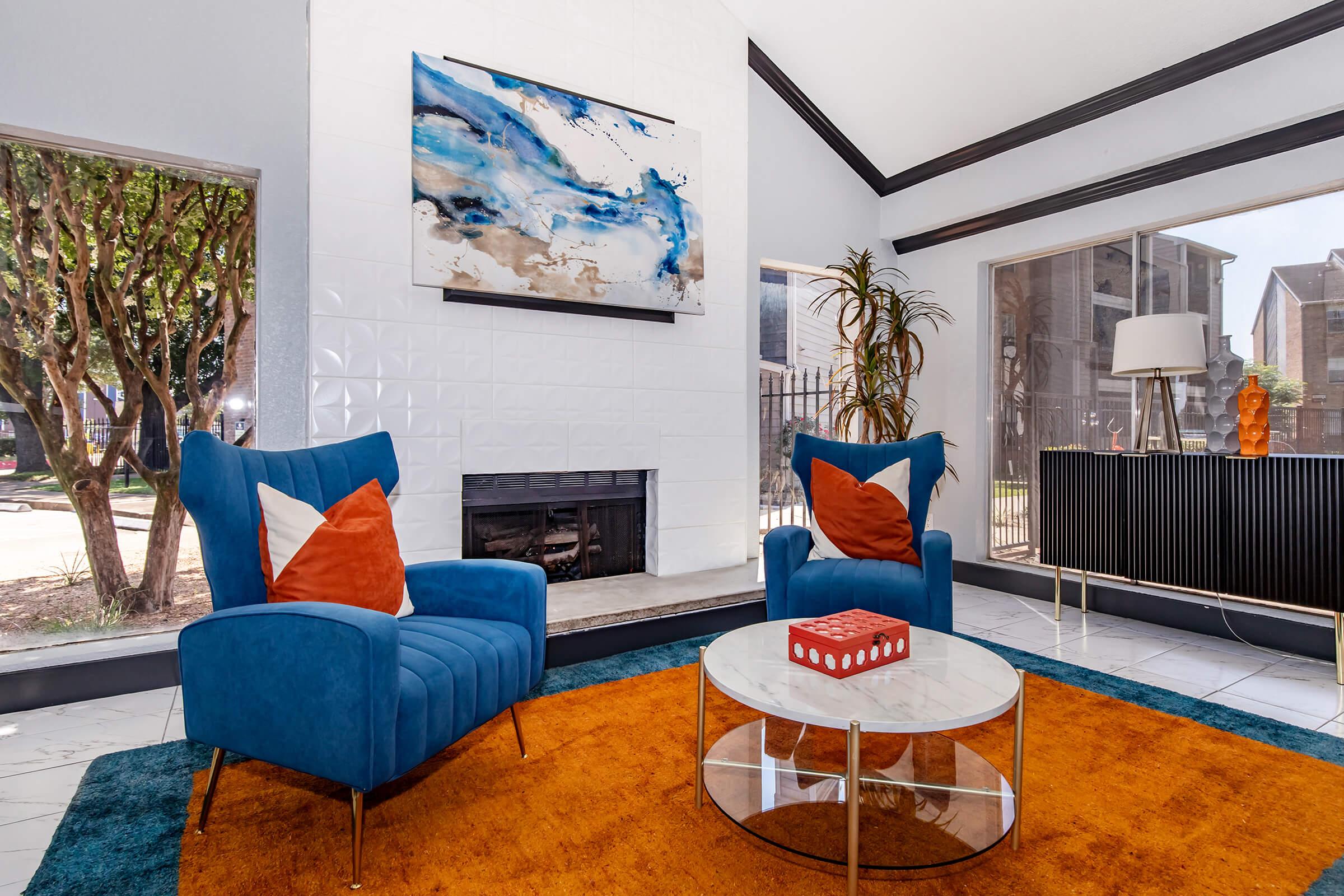 A cozy living room featuring two blue chairs with orange cushions, a round glass coffee table, and a vibrant orange rug. A fireplace is situated against a white tiled wall, and a colorful abstract painting hangs above. Large windows let in natural light, revealing a garden view outside.