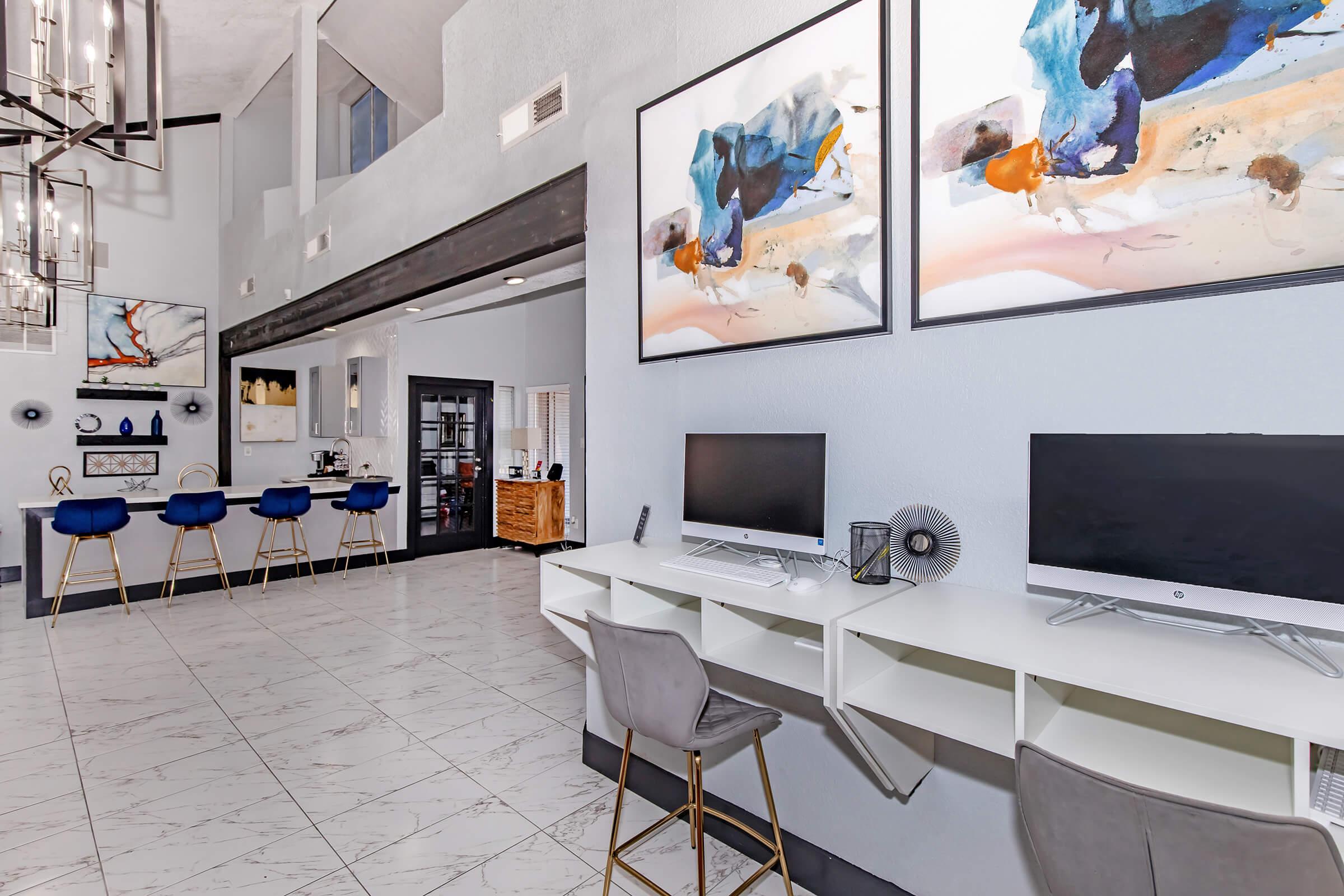 A modern workspace featuring two desktop computers on a white desk, stylish gray chairs, and contemporary artwork on the walls. In the background, there's a bar area with blue stools and decorative shelves, along with elegant lighting fixtures, creating a sophisticated and inviting atmosphere.