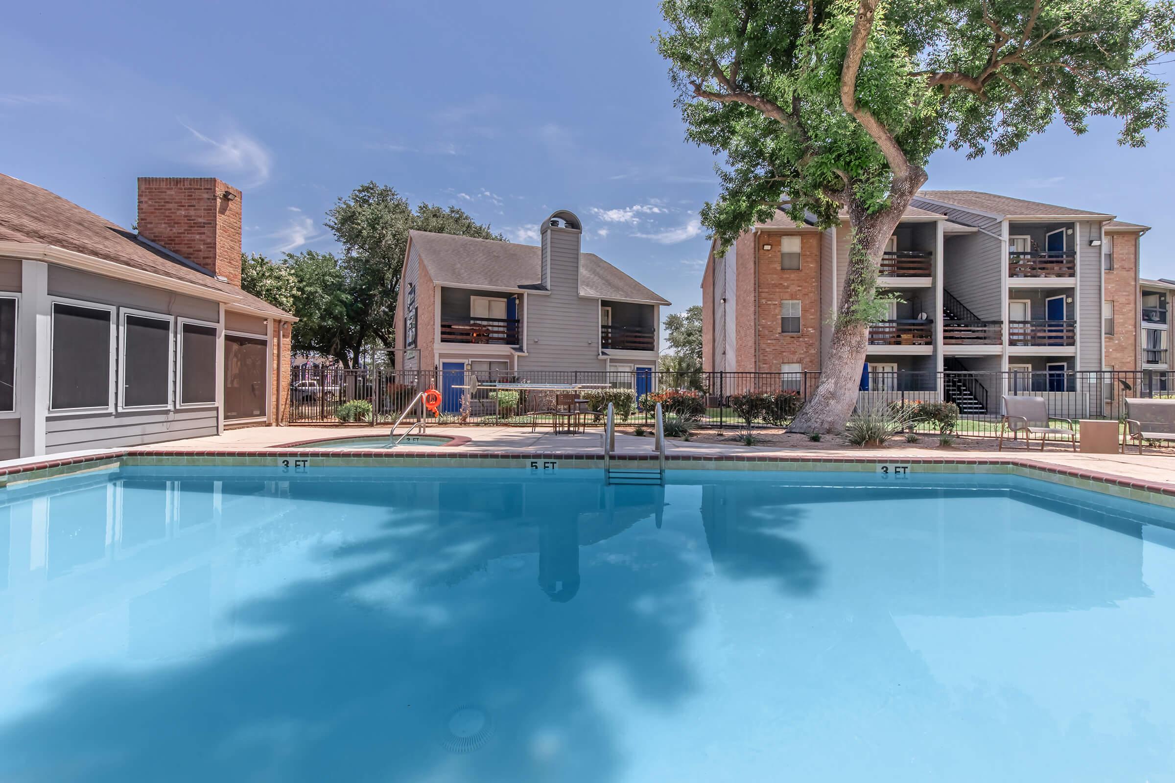 A clear blue swimming pool surrounded by well-maintained landscaping. Apartment buildings in the background feature balconies and outdoor seating areas. There is a large tree providing shade near the pool, along with a lounge chair and a poolside safety buoy visible. The sky is sunny with a few clouds.