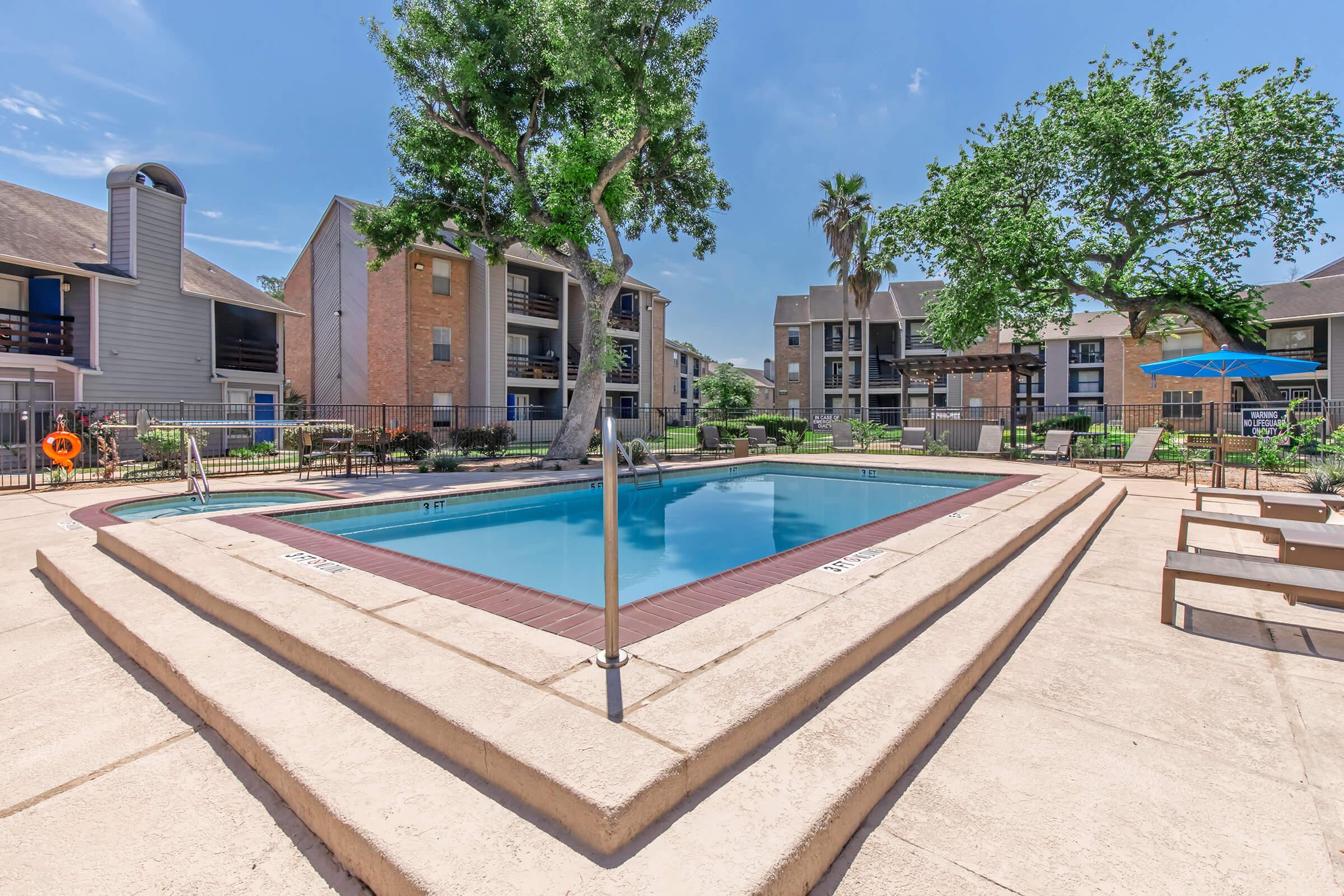 A clear blue swimming pool surrounded by a spacious deck, palm trees, and green lawns. Nearby, there are residential buildings with balconies. Sun umbrellas and lounge chairs are positioned around the pool area, creating a relaxing outdoor atmosphere on a sunny day.