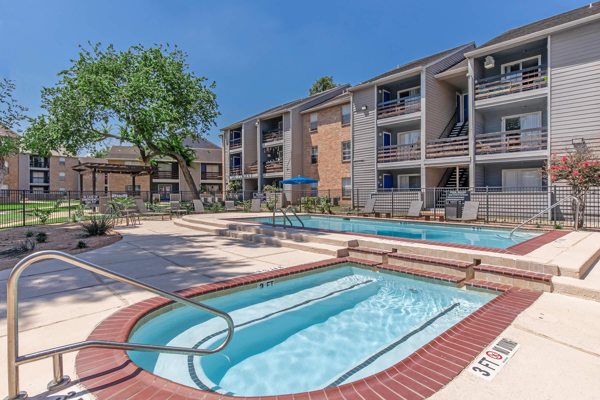 A sunny pool area featuring two hot tubs and a swimming pool surrounded by a concrete deck. Lush green trees and landscaping enhance the space, with buildings in the background providing residential units. Lounging chairs are placed near the water, creating a relaxing atmosphere.