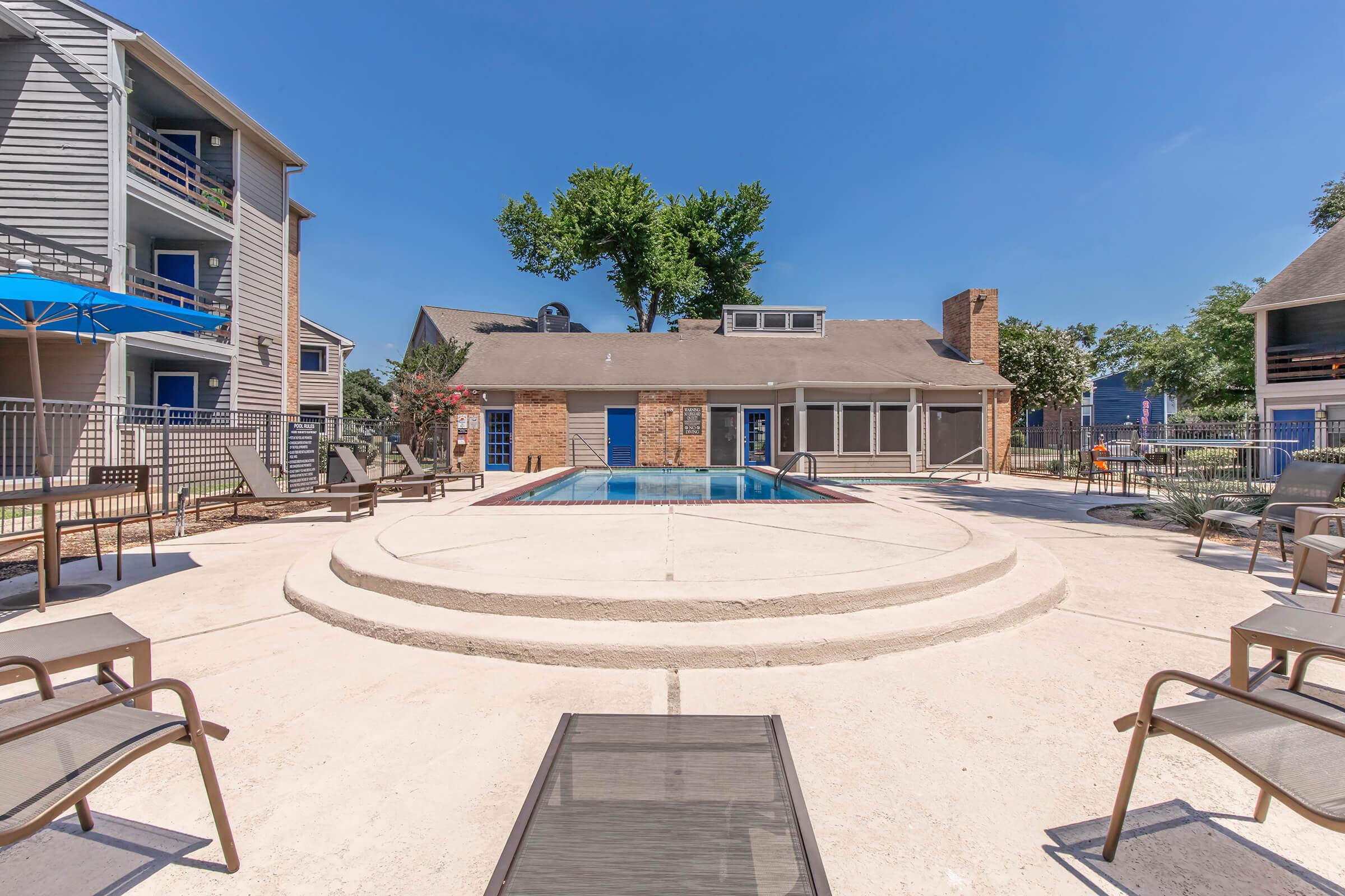 Swimming pool area with a circular deck surrounded by lounge chairs. A building with blue doors and large windows is located in the background. Clear blue sky and trees are visible, creating a sunny and inviting atmosphere.