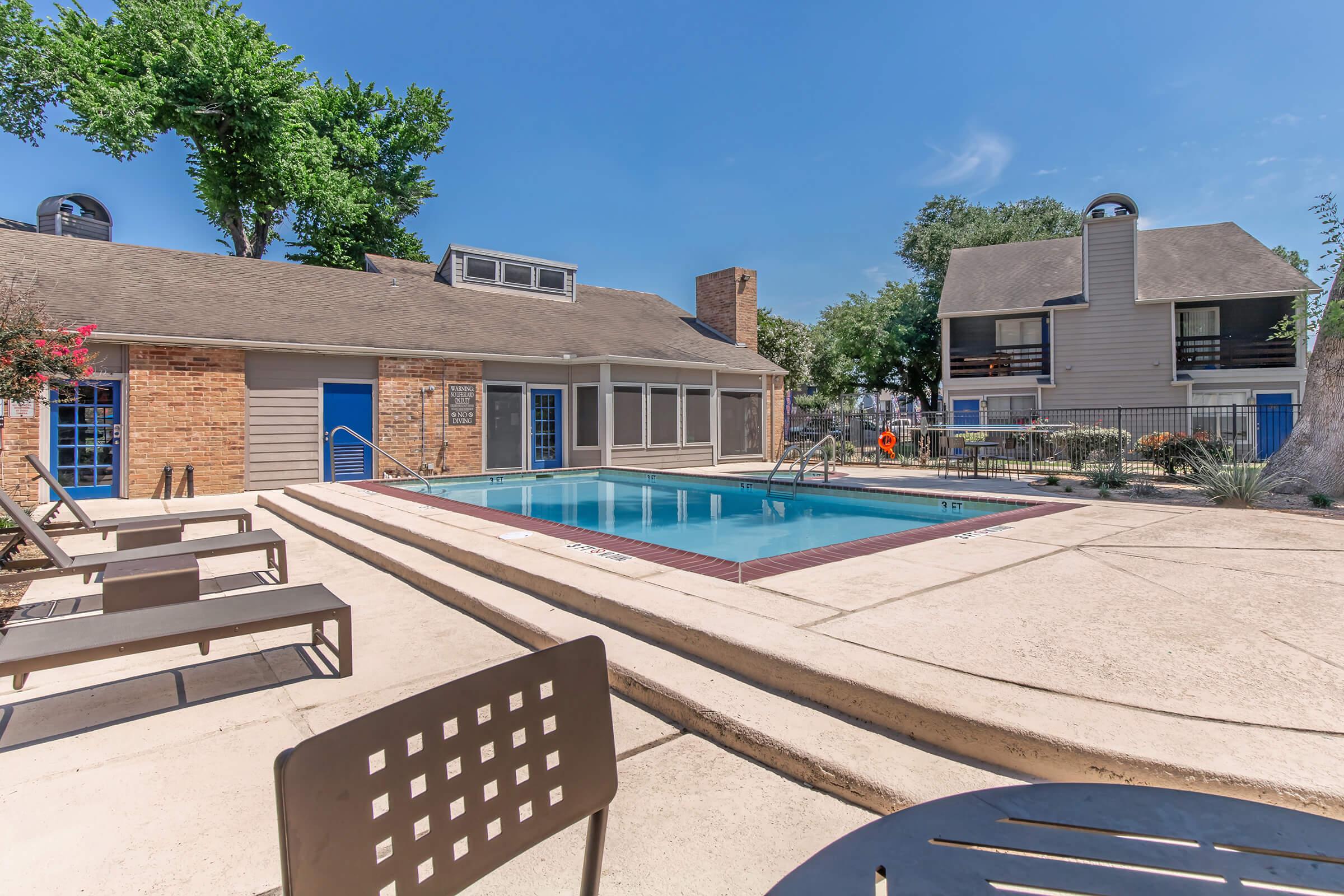 A swimming pool surrounded by lounge chairs and a patio area, with a brick building to one side and residential units in the background. The scene is set in a sunny outdoor environment with trees in the vicinity, creating a relaxing atmosphere.