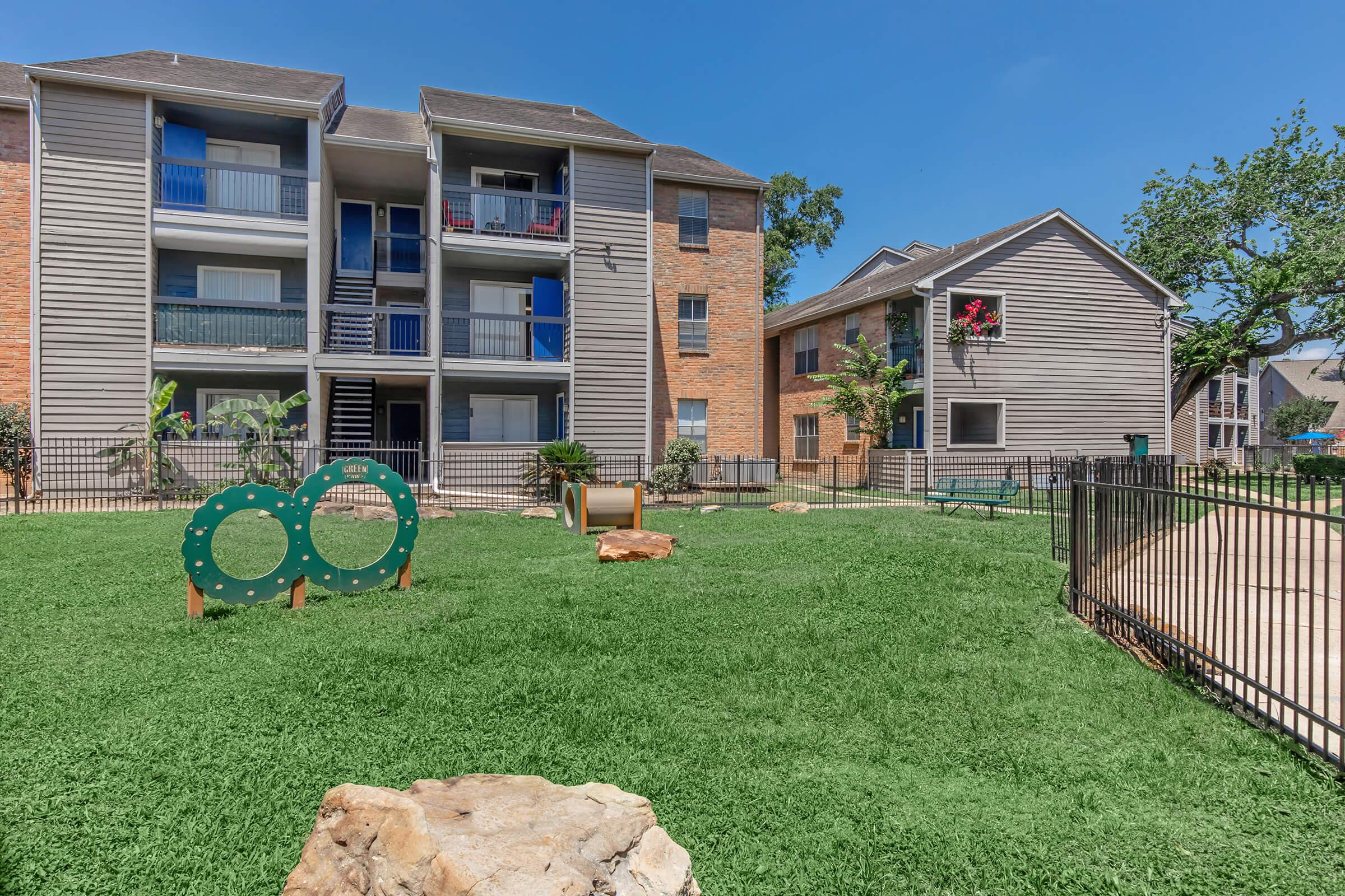 A well-maintained apartment complex featuring two buildings surrounded by lush green lawns. In the foreground, there are playful structures for children, including a tunnel and a bench, along with rocks integrated into the landscape. The sky is bright blue, creating a cheerful atmosphere.