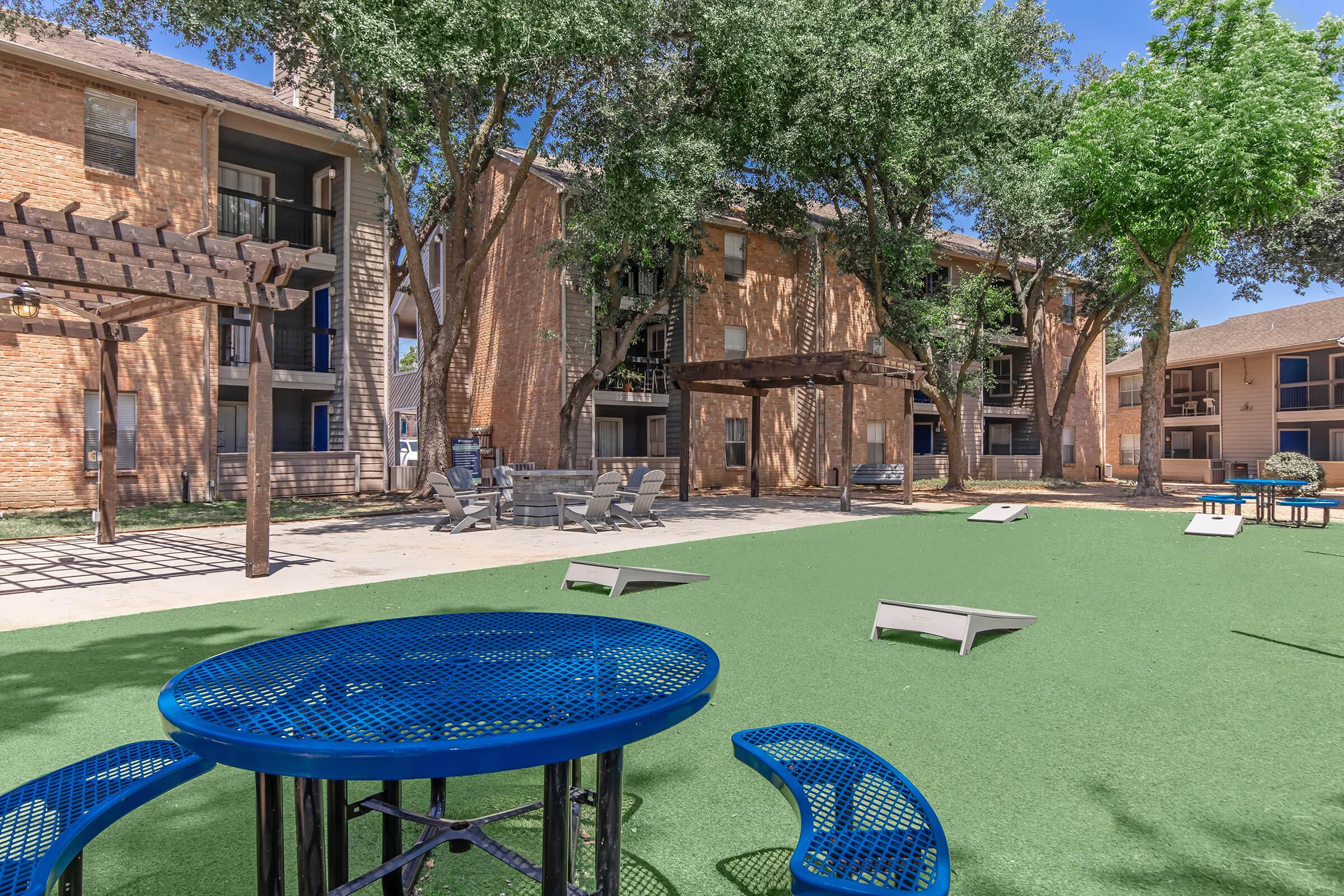 A landscaped courtyard featuring artificial grass, a blue picnic table with attached benches, and several lounge chairs. In the background, there are two apartment buildings surrounded by trees, with a pergola structure and cornhole boards set up for games.