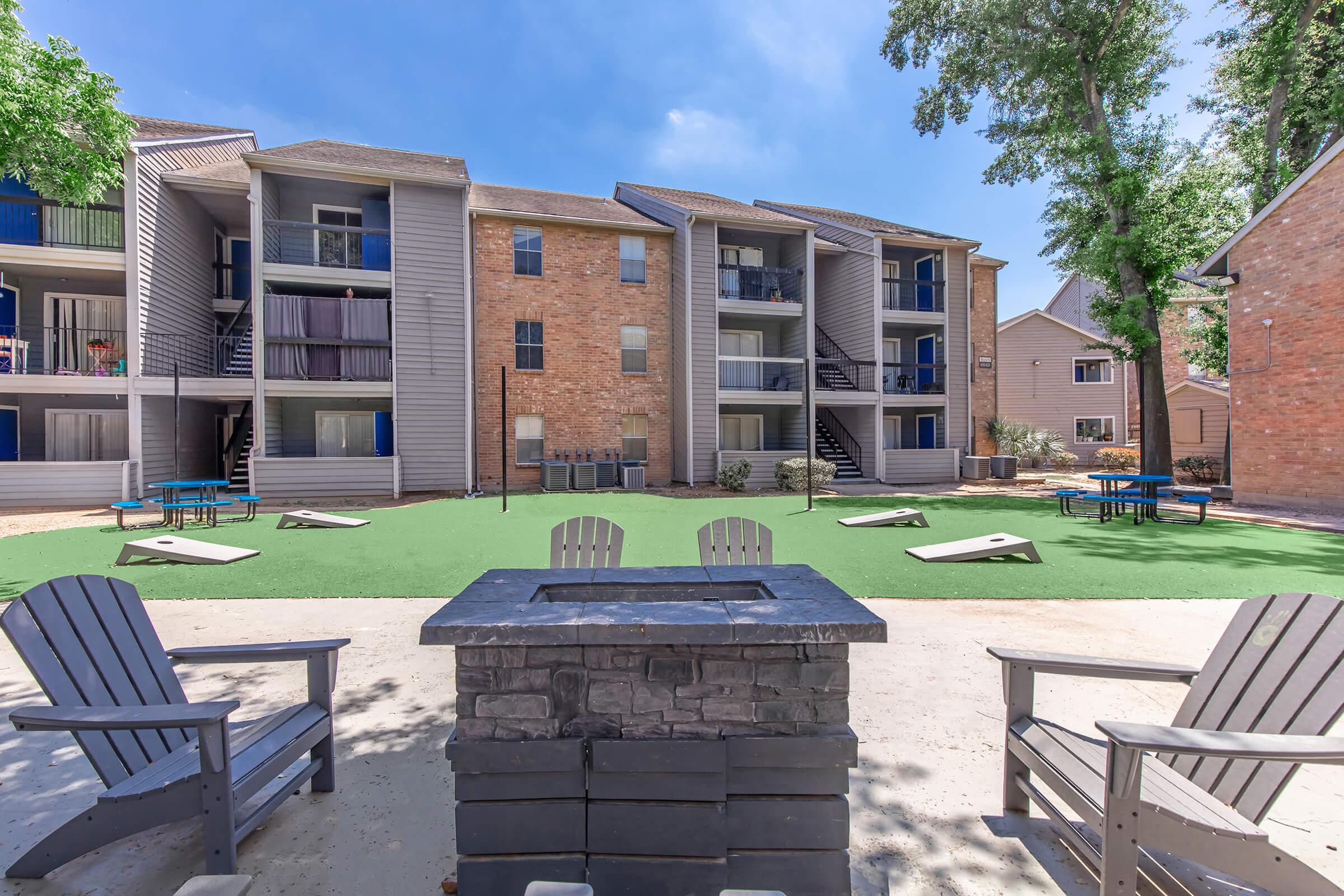 A communal courtyard featuring a green lawn area surrounded by apartment buildings. In the foreground, there are two Adirondack chairs facing a stone fire pit. A few picnic tables and bean bag toss games are visible in the background, with trees providing shade under a clear blue sky.