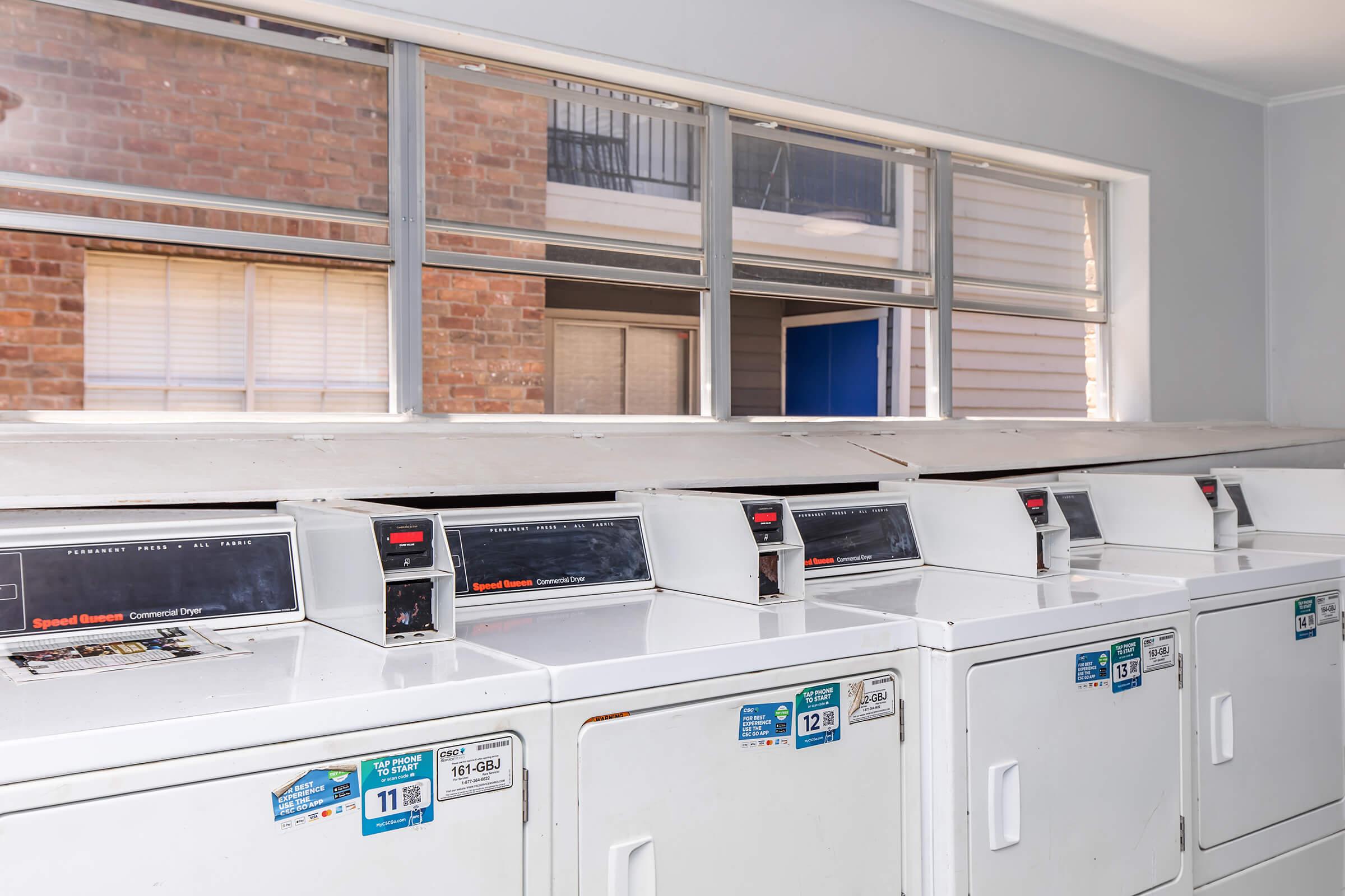 A row of white commercial washing machines and dryers in a laundry room. Large windows provide natural light, with a view of a brick exterior and nearby buildings. Various labels and prices are visible on the machines.