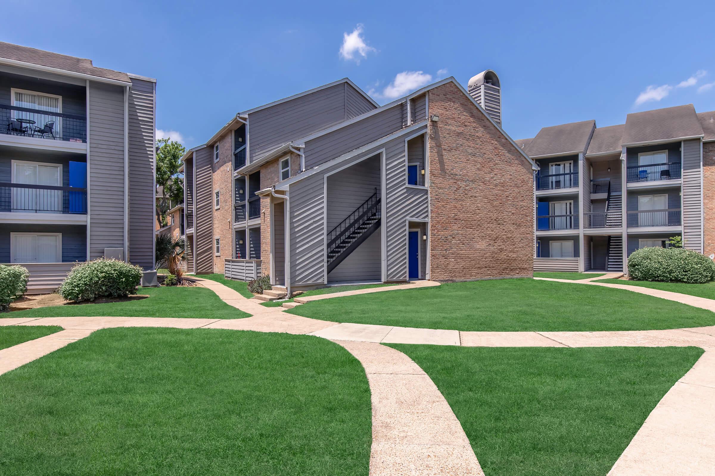 A residential apartment complex featuring two buildings with a mix of brick and siding exteriors. The landscape includes well-maintained green lawns and a paved walkway connecting the buildings. The sky is clear with a few clouds, suggesting a bright day.