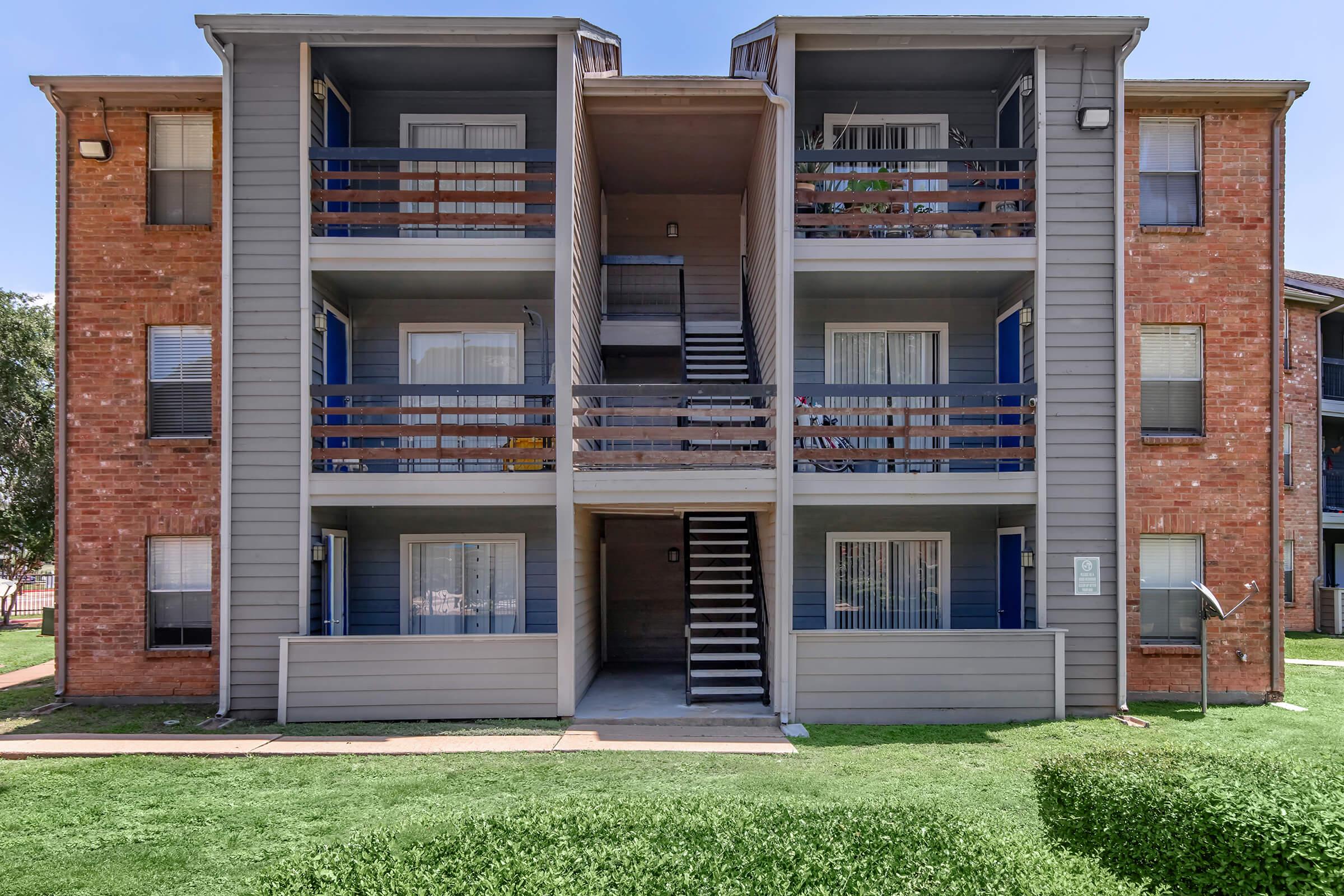 A multi-story residential building featuring brick and gray siding, with four balconies and a staircase in the center. The surrounding area has green lawn and trees, and there are several windows with vertical blinds visible.