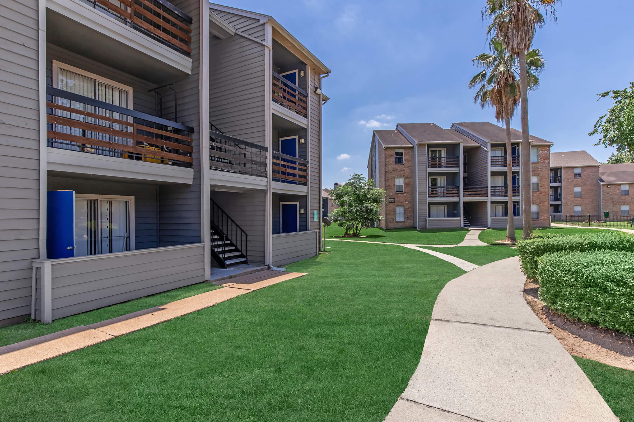 Two multi-story apartment buildings are surrounded by well-maintained green lawns and palm trees. A curved concrete pathway leads through the grass, connecting the buildings, which feature balconies and outdoor stairs. The sky is clear and blue, indicating a sunny day.