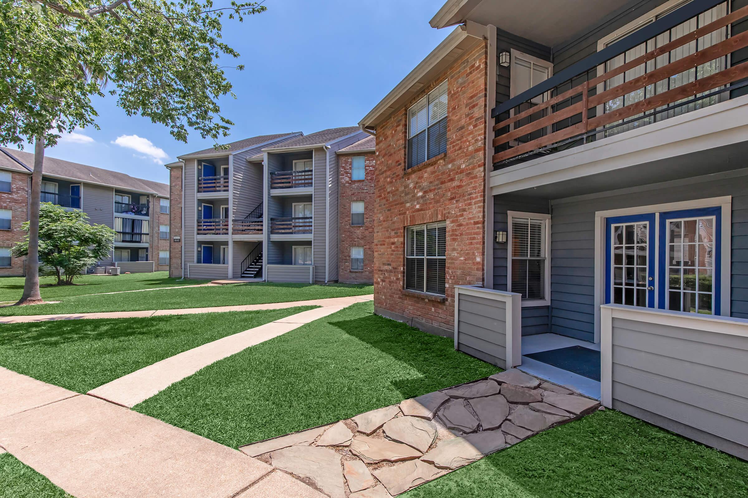 A view of a well-maintained apartment complex featuring several buildings with a mix of brick and exterior siding. Lush green lawns and pathways connect the buildings, with trees providing shade. The image captures a sunny day, highlighting the inviting outdoor space.