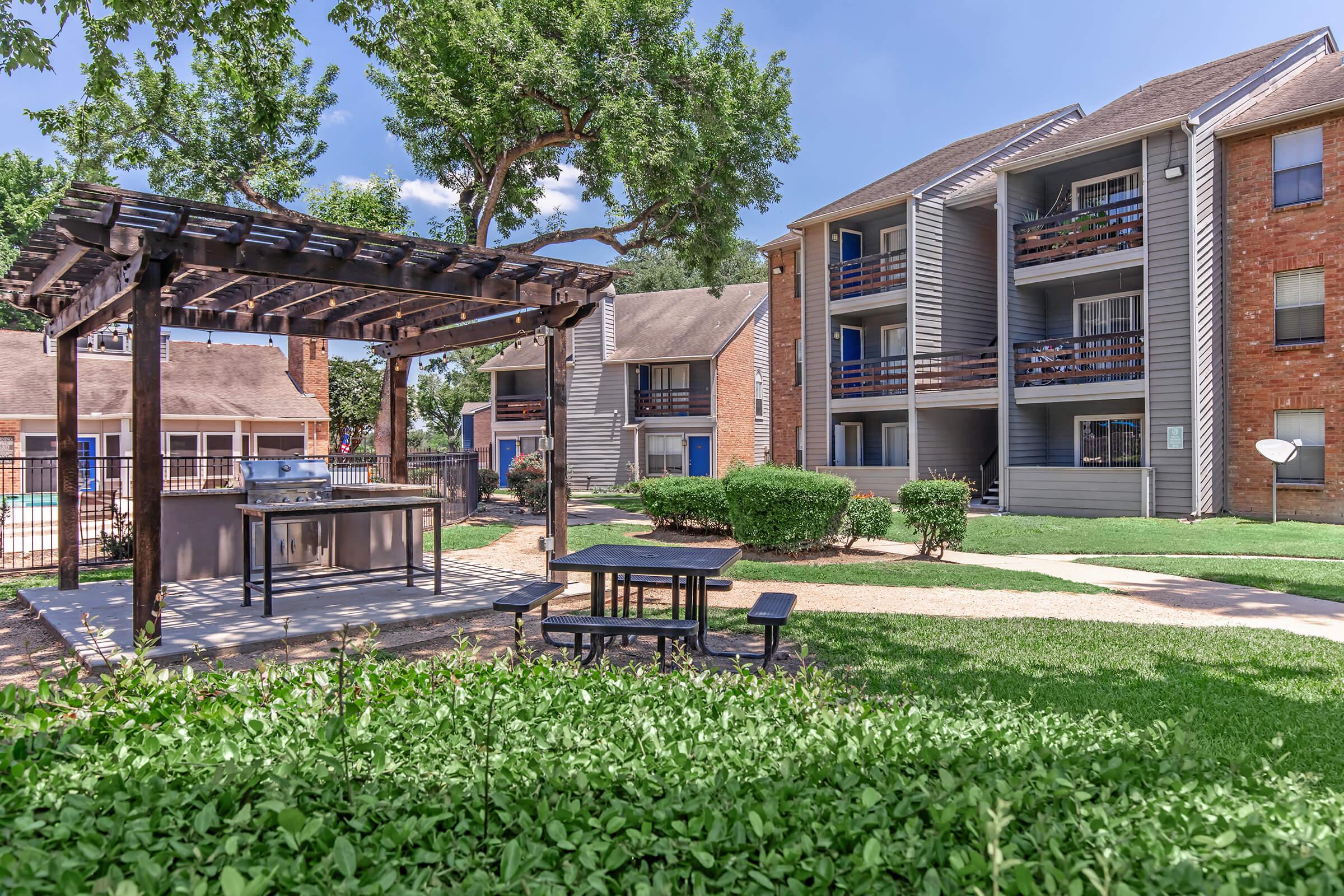 A landscaped area featuring a picnic table and grill under a wooden pergola, surrounded by green bushes. In the background, there are two multi-story apartment buildings with balconies, set against a clear blue sky. The scene conveys a welcoming outdoor communal space.
