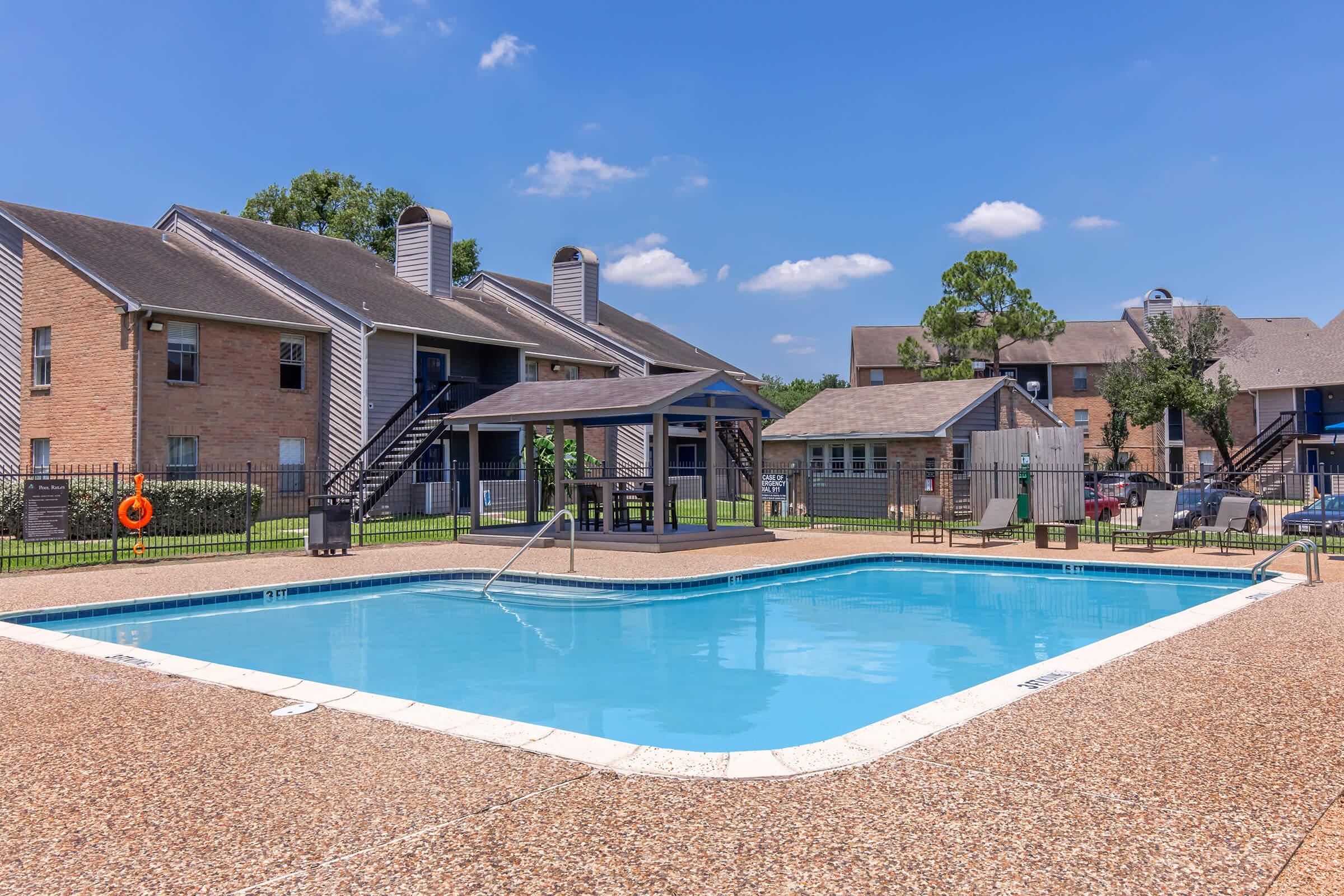 A clear blue swimming pool surrounded by a tiled deck, with a gazebo nearby. In the background, there are two multi-story apartment buildings and a well-maintained lawn. The sky is bright with a few clouds, creating a sunny, inviting atmosphere.