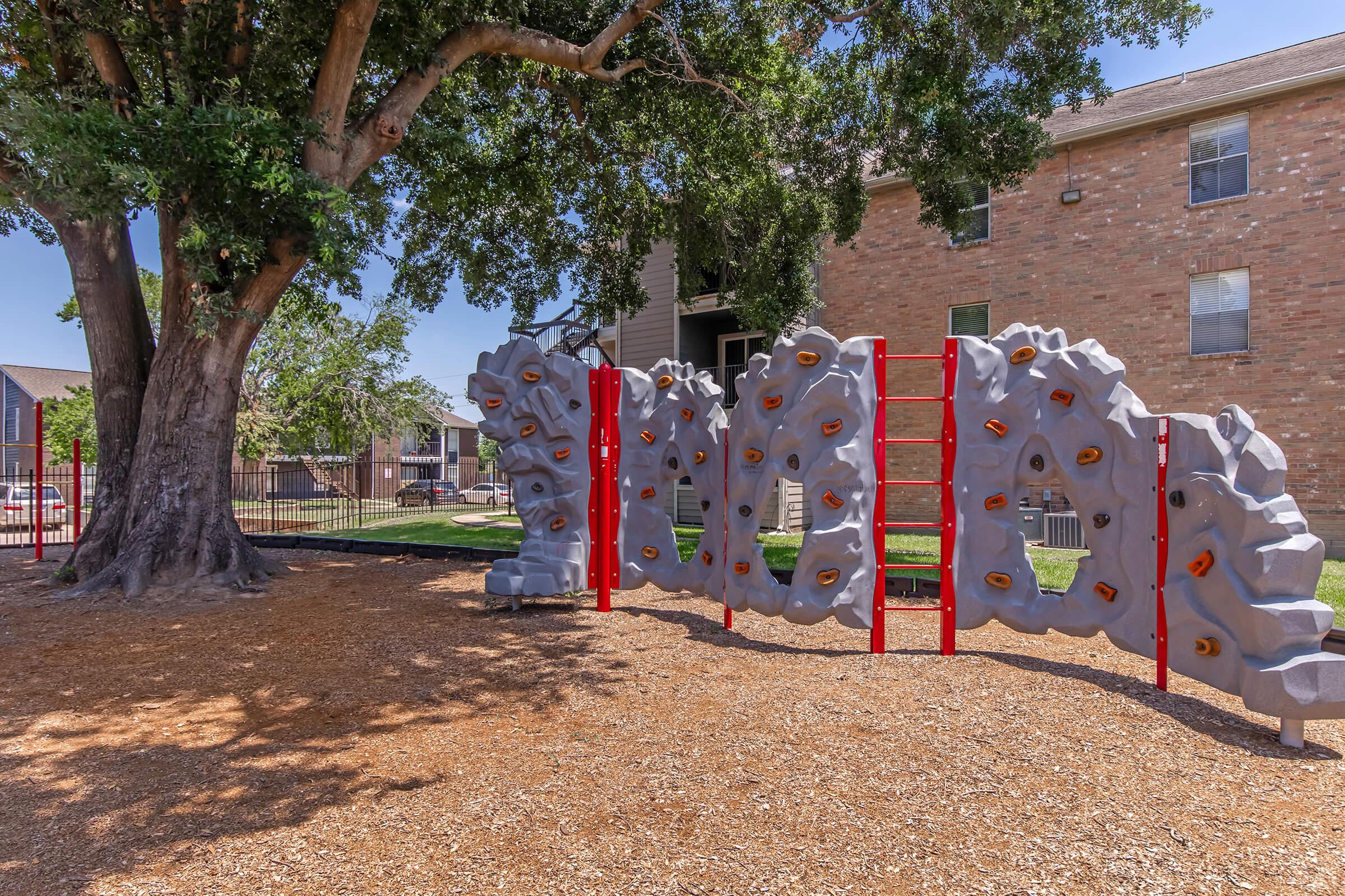 A children's climbing structure made of gray rock-like panels with orange handholds, situated in a playground area with a tree and an apartment building in the background. The ground is covered with wood chips, and there are red climbing ladders attached to the structure.