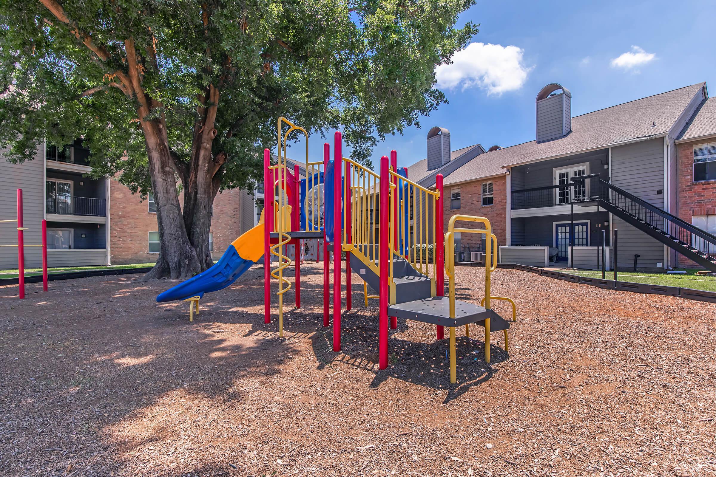 Colorful playground equipment with a slide, climbing structure, and steps, surrounded by mulch and trees. In the background, there are residential buildings with balconies and blue sky.