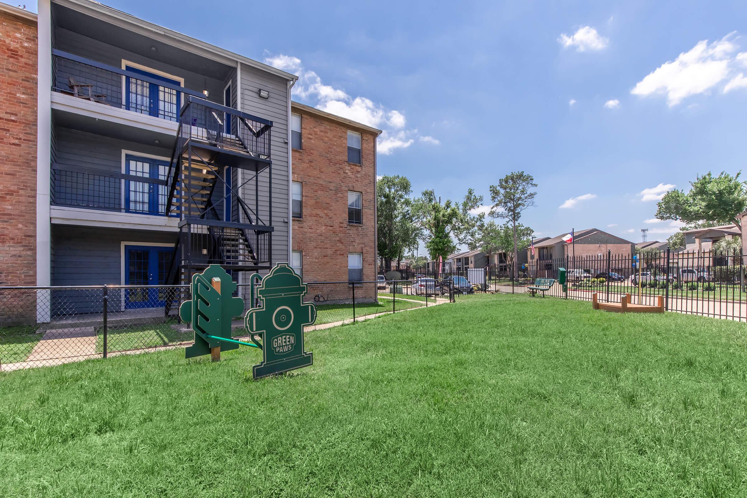 A view of a residential apartment complex featuring a green lawn with two decorative signs: one resembling a tree and the other a fire hydrant. In the background, a three-story building with a staircase is visible, along with additional buildings and trees under a clear blue sky.