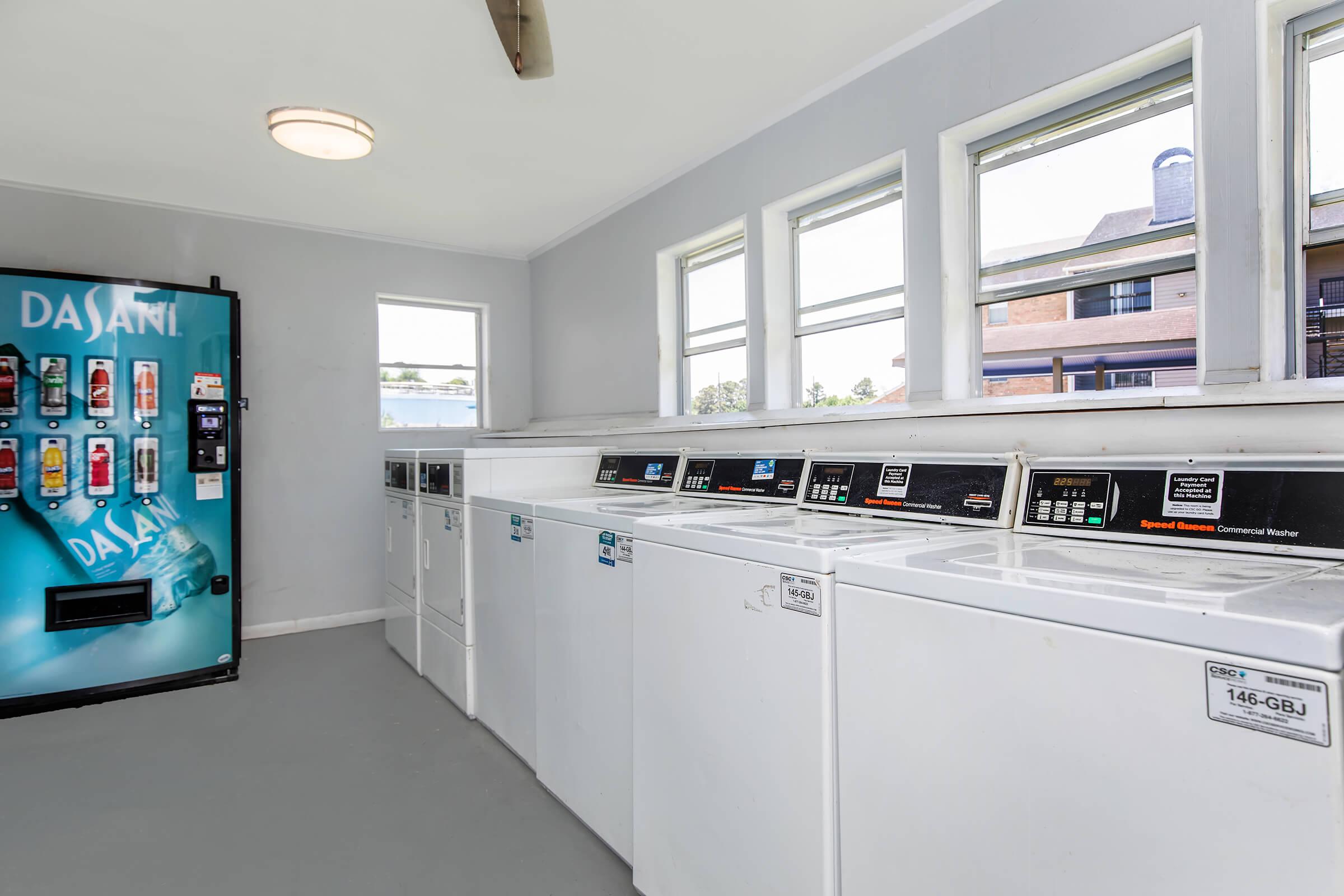 A clean laundry room featuring several white washers and dryers in a row. Large windows allow natural light to fill the space. On one side, there's a vending machine stocked with beverages, including Dasani water, adding convenience for users. The walls are painted in a light gray color.