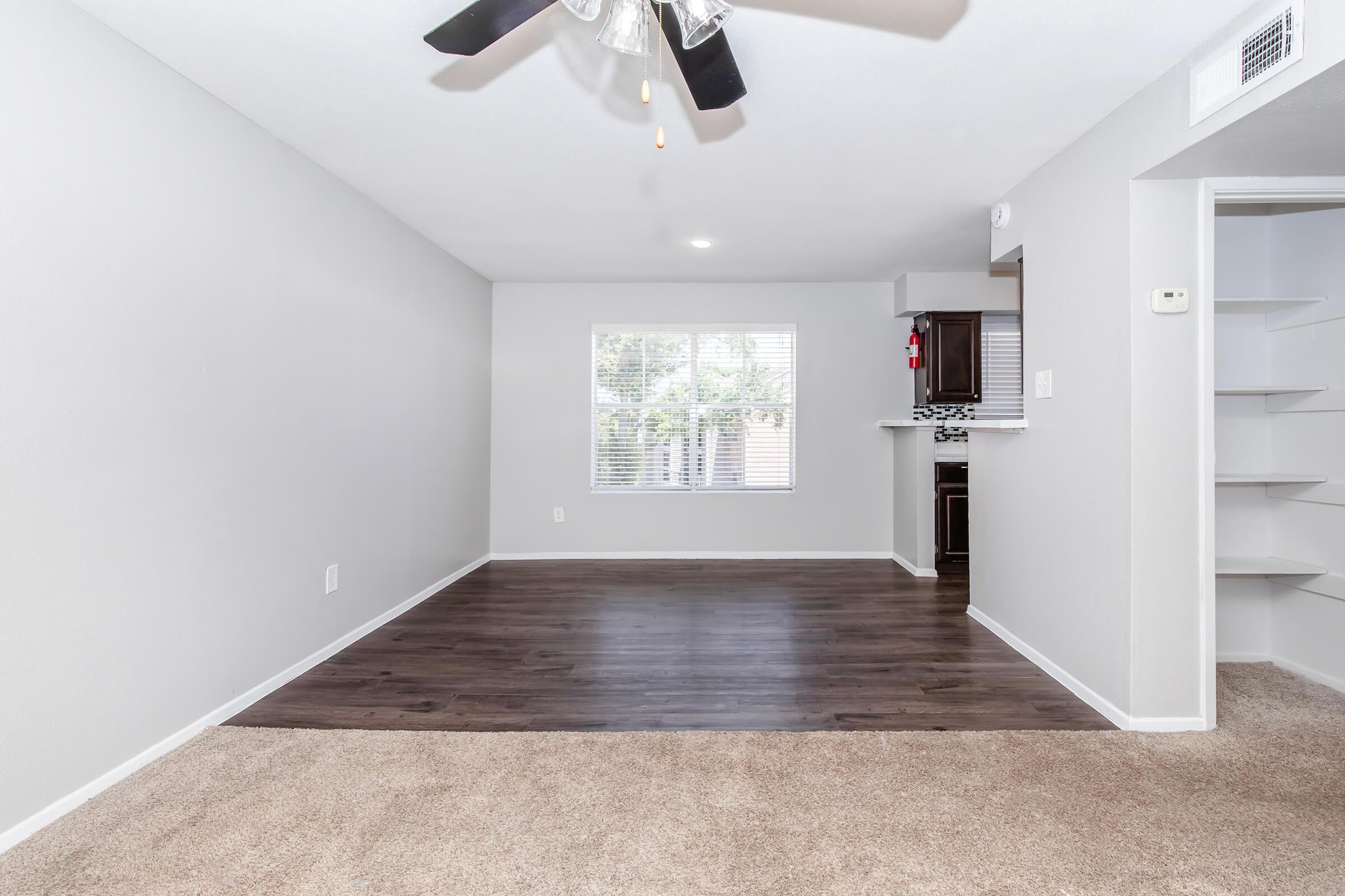 A spacious living area with light gray walls and a ceiling fan, featuring hardwood flooring. A large window allows natural light to enter, and an open layout leads to a kitchen area in the background. There's a small built-in shelf on the right side. The room is inviting and freshly decorated.