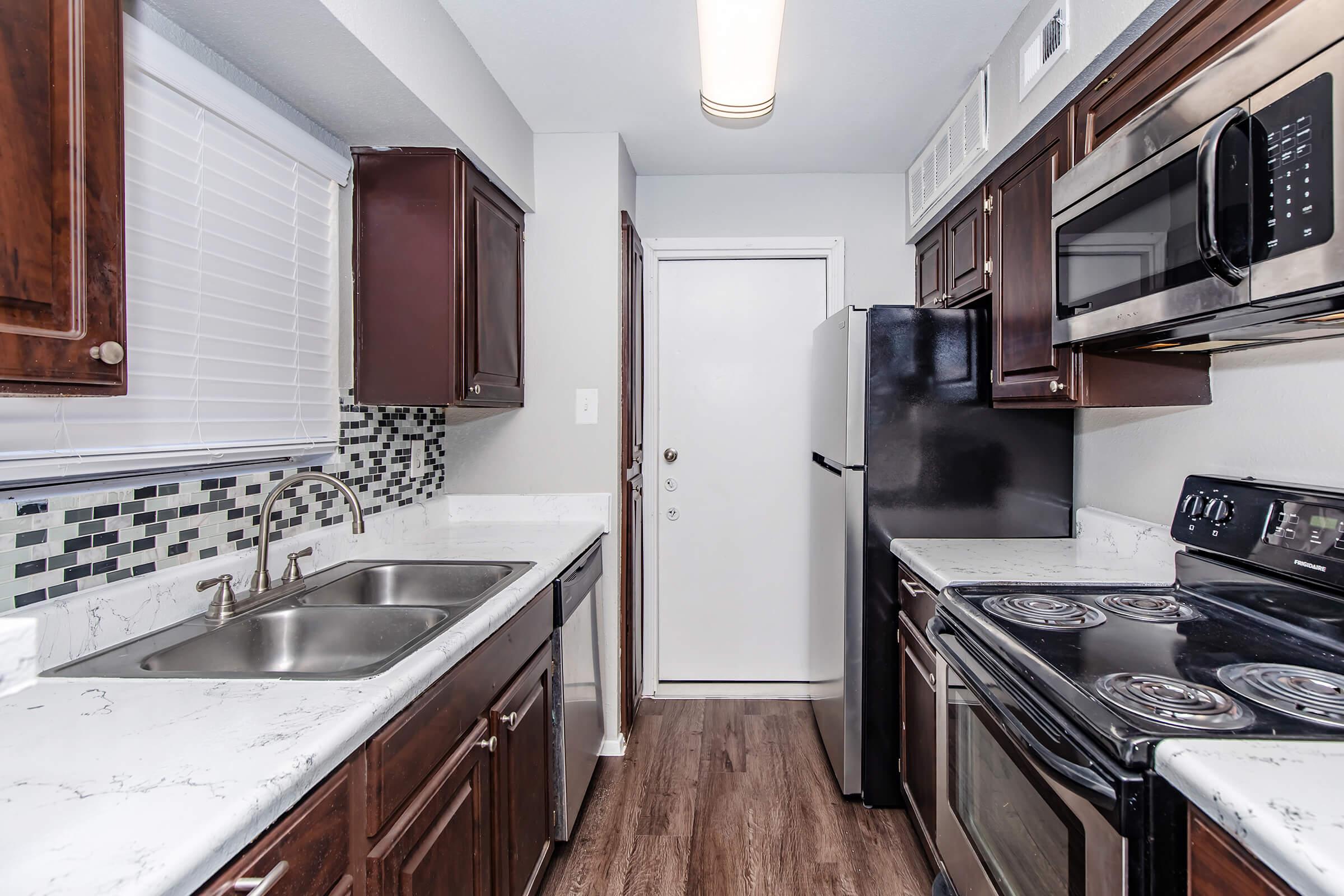 Modern kitchen featuring dark wooden cabinetry, a double sink, and granite countertops. Stainless steel microwave and stove with an adjacent black refrigerator. Includes a tiled backsplash and a door leading outside, with a window allowing natural light.
