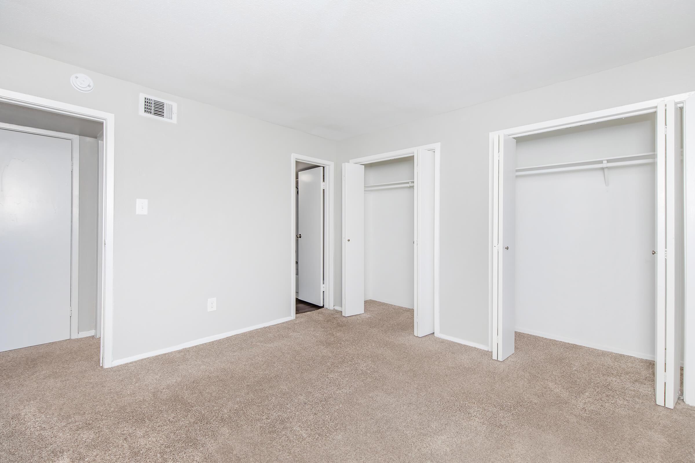 A spacious, empty room with beige carpeting and light gray walls. There are two open closet doors on the left and one door in the back. The overall atmosphere is bright and minimalistic, suitable for various uses.