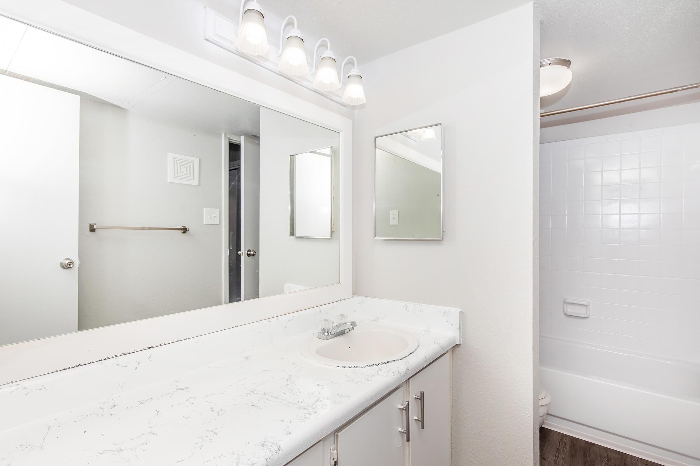 A modern bathroom featuring a light-colored countertop with a sink, two mirrors on the wall, and a shower area with a bath. The walls are painted in neutral tones, and there is ample natural light from a window. A towel rack is mounted beside the door.